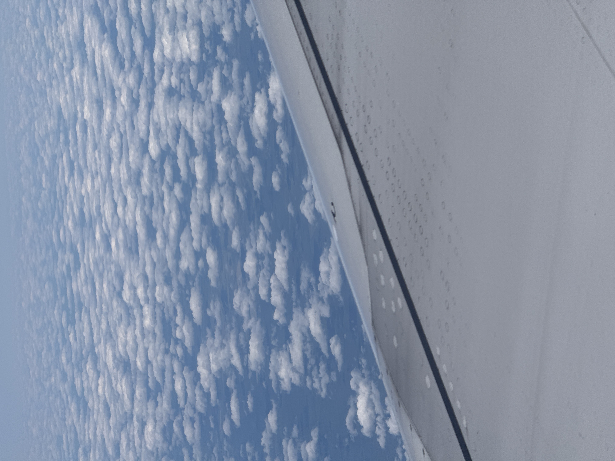 The image shows a view from an airplane window, looking down at a vast expanse of white clouds spread across a blue sky. The clouds appear to be at a low altitude, forming a textured, undulating layer. In the foreground, a portion of the airplane's wing is visible, its metallic surface showing a pattern of small circular indentations and a dark, straight line running diagonally across it. The overall scene is one of serene aerial perspective, with no discernible elements that would allow for specific location identification within Kazan, Russia, or any other city.