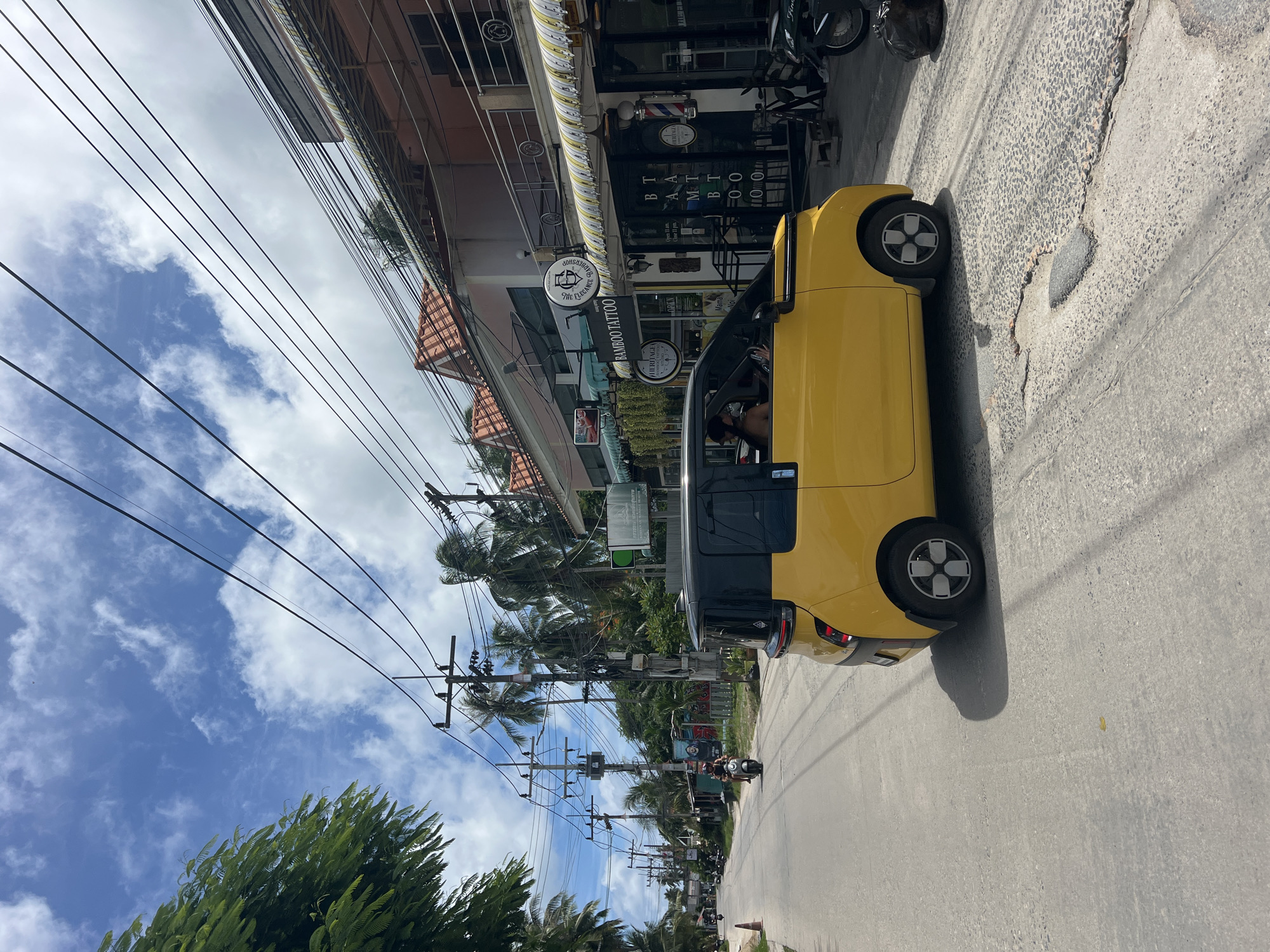 The image depicts a street scene in Tambon Ko Pha Ngan, Thailand, characterized by a bright yellow car with a black roof driving on the right side of the road. The street is paved and appears somewhat worn, with visible patches and cracks. On the right side of the road, there are several shops, including one with signage indicating "BAMBOO TATTOO" and another with "HERITAGE." Some of these shops have awnings and outdoor seating. Power lines crisscross above the street, supported by utility poles. Palm trees line the side of the road, and lush green foliage is visible in the background. The sky is blue with scattered white clouds. In the distance, more buildings and trees are visible, as well as other vehicles on the road, including a scooter carrying two people. A person is visible inside the yellow car, appearing to be driving. The overall impression is a casual, tropical street environment.