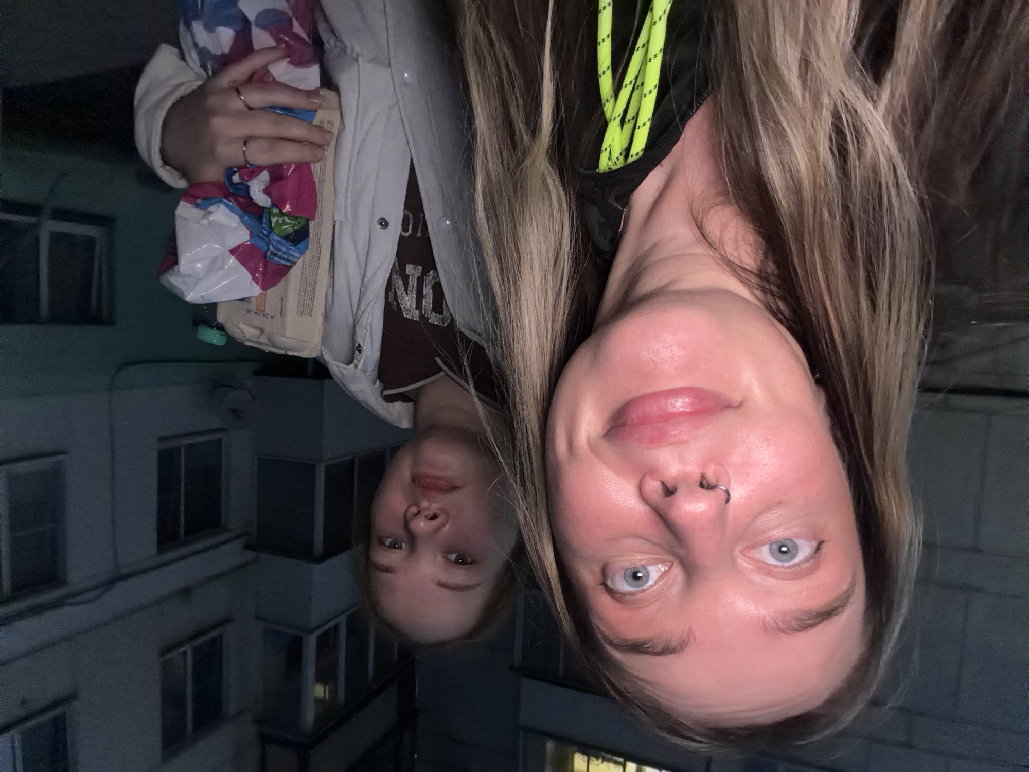 Two women are pictured from the neck up, seemingly reflected in a dark surface. One woman, closer to the viewer, has light skin, blonde hair, and blue eyes. She has a silver ring in her nose. The second woman is visible behind and to the left, with similar coloring. Both are partially obscured by the angle. In the background, there is a building with multiple windows, suggestive of an urban environment. The reflection suggests the image was taken at night. In the upper left corner, someone is holding a bag of groceries and some other objects. Based on the location hint, this is likely a scene in Chaikovsky, Russia.