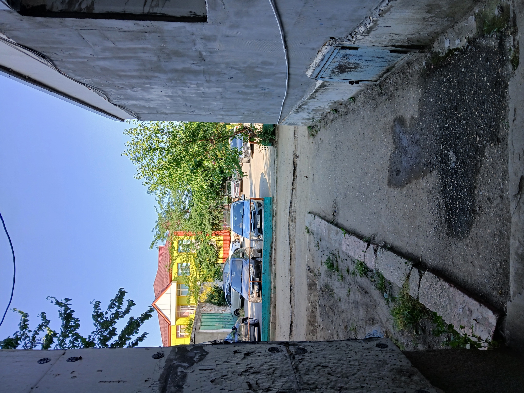 The image depicts an outdoor scene in Makhachkala, Russia. In the foreground, a rough concrete structure frames the view. The scene opens to a bright, clear sky, suggesting it is daytime with fair weather.
The main subjects are several cars parked in a row. One car, a dark sedan with a license plate visible, is in the mid-ground. Behind it, another car, also a dark sedan, is parked. Further back, a blue car is visible, and there might be other vehicles obscured by trees. These cars are parked on a paved area that transitions to a dirt and gravel path on the right side of the image.
To the left, a vibrant yellow building with a red-tiled roof is partially visible, featuring multiple windows. Lush green trees with dense foliage are scattered throughout the scene, providing a contrast to the urban elements. A dark metal gate is also visible in front of the building.
On the right side of the image, the ground is a mix of concrete and gravel. A low stone border runs along the edge of the paved area, with patches of grass and weeds growing in the crevices. A blue metal utility box or hatch is set into the concrete wall. The overall impression is one of a residential or communal outdoor space, possibly a courtyard or alleyway, in a sunny, possibly warm, climate. There are no people visible in the image. The visible license plate on the dark sedan appears to have Cyrillic characters.