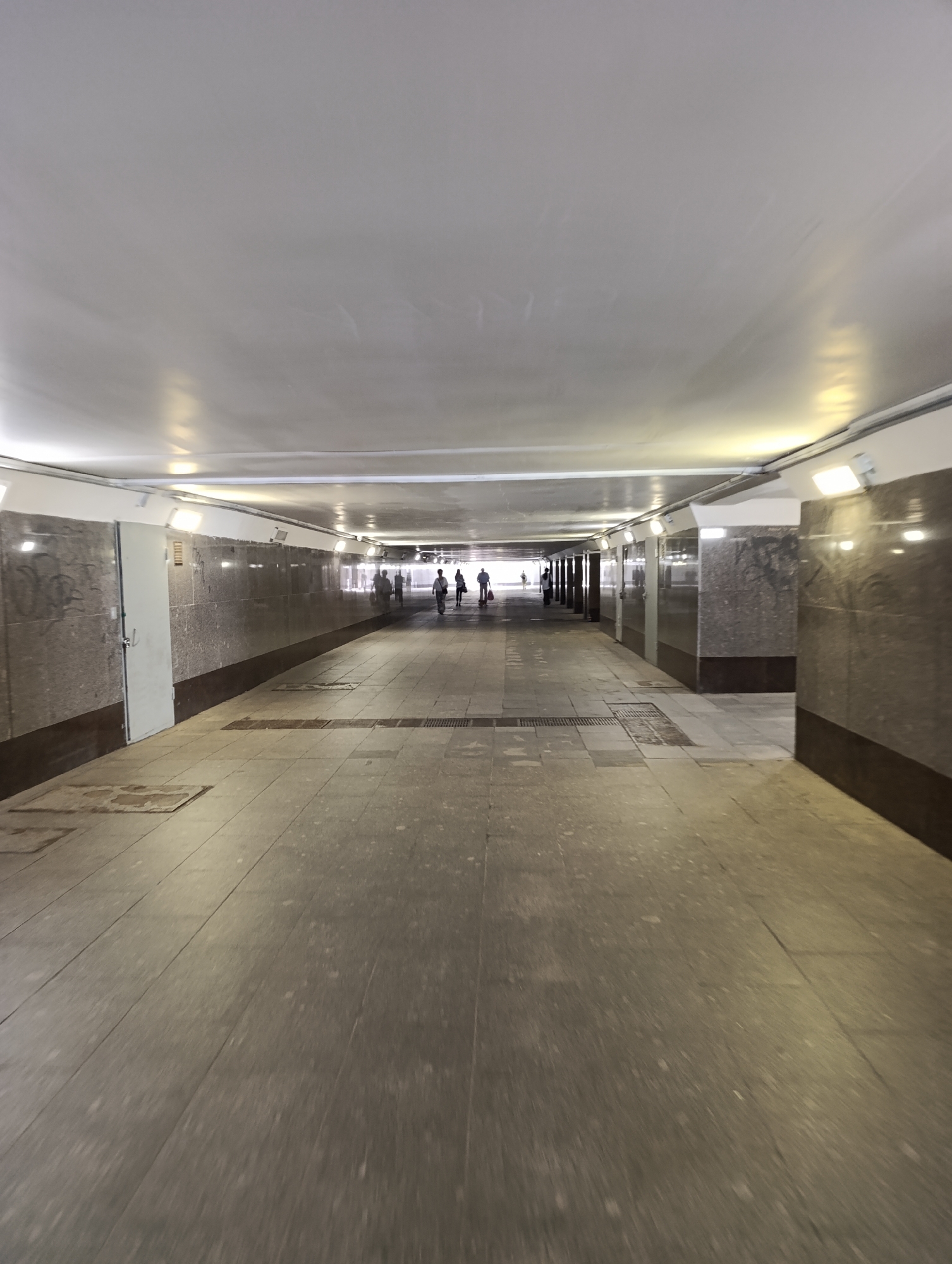 The image depicts a long, well-lit underground pedestrian tunnel, likely an underpass in Moscow, Russia. The perspective is from inside the tunnel, looking towards its distant exit.
The tunnel features a flat, light-colored ceiling, possibly made of a stretched or panel system, which reflects the numerous bright, rectangular lights mounted along the upper walls. These lights illuminate the entire passageway. The upper sections of the walls are also light-colored, while the lower half is clad in dark-patterned, possibly granite or marble-like, stone panels. A white door is visible on the left wall, near the foreground, with some faint, dark graffiti on the adjacent stone panels. Further down the tunnel, similar paneling and structural elements are repeated on both sides.
The floor is paved with large, rectangular grey or light brown tiles, some of which appear worn or stained, with visible grates or drainage covers in the middle.
In the far distance, several indistinct human figures are visible, silhouetted against the brighter light emanating from the tunnel's end. They appear to be walking through the underpass. One figure on the left side in the distance seems to be pushing a cart or similar object. The overall atmosphere is functional and utilitarian. No specific time of day is discernible from the internal lighting, but the light at the exit suggests it might be daytime outside. There is no legible text apart from the faint, unidentifiable graffiti.