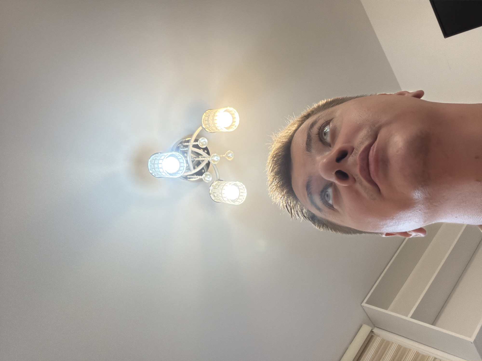 A low-angle, head-and-shoulders shot captures a person from the chest up, looking towards a ceiling fixture with multiple lights. The individual's face is mostly in focus, with eyes directed upward. The ceiling is white and shows a chandelier with several crystal-like shades and exposed bulbs, casting a warm light. The background consists of a white ceiling and a hint of shelving units or a built-in cabinet to the left. The image does not provide information about the city of Budva, Montenegro.
