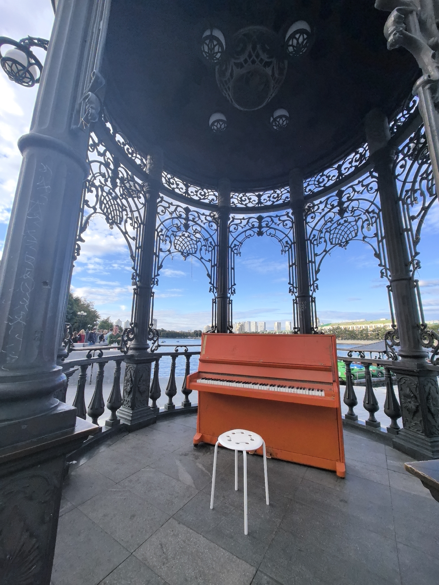The image shows an orange upright piano and a white stool on a paved area within a decorative gazebo. The gazebo has dark metal ornate railings and columns, with a view of a body of water and buildings in the background. In the distance, several people are visible walking along a waterfront. The sky is blue with scattered clouds. The piano and stool are positioned centrally within the gazebo. The architecture of the gazebo suggests it is a public space, possibly a bandstand or a decorative feature in a park. The style of the buildings and the water body are consistent with urban waterfronts. This scene is located at the plot of land near the monument to Vladimir Mayakovsky on the embankment of the Iset River in Yekaterinburg, Russia.
