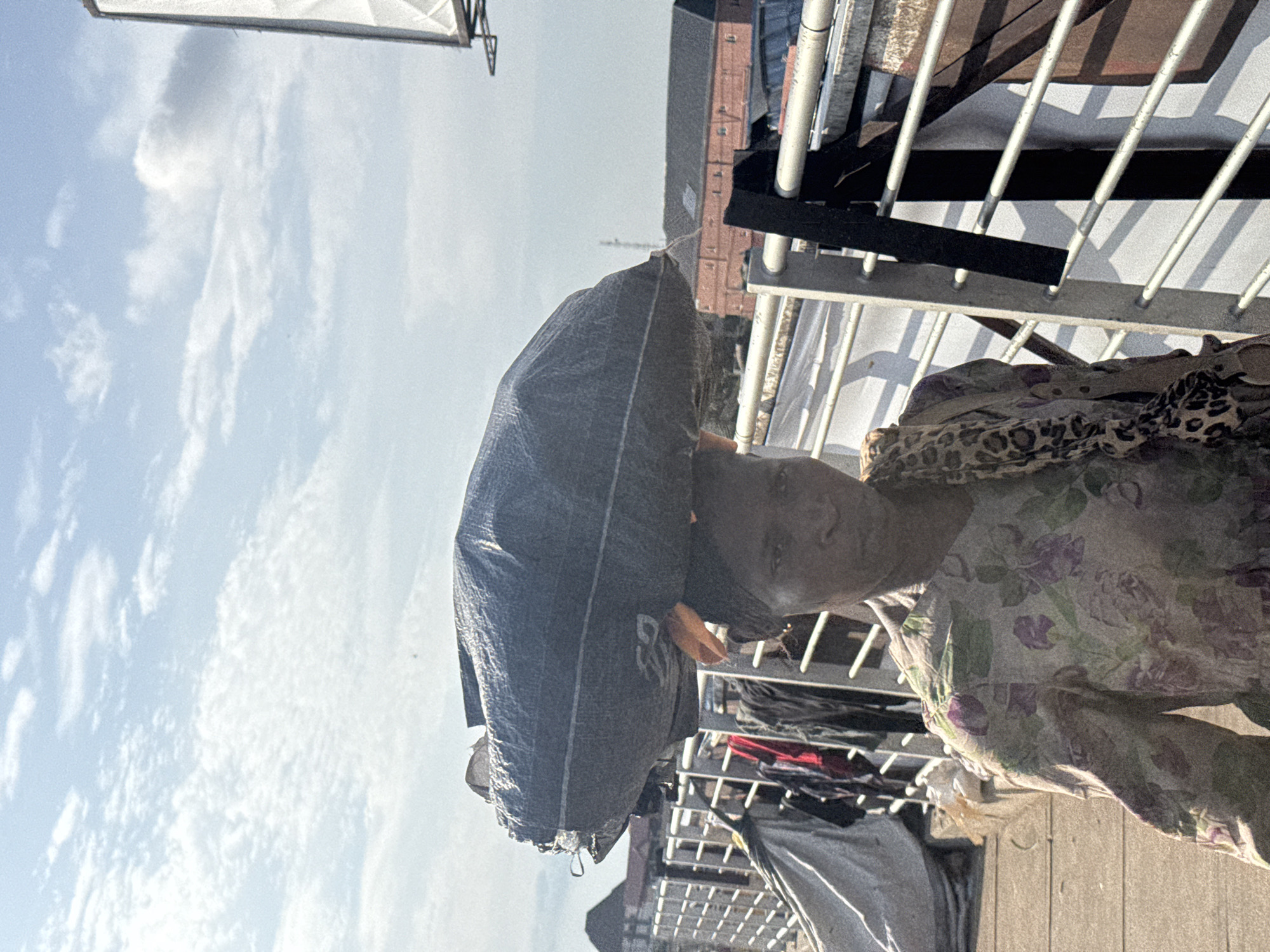 A woman carries a large, dark-colored bundle on her head in a city environment, likely Onitsha, Nigeria. She is seen from the chest up, facing the camera with a neutral expression. The bundle is wrapped in plastic sheeting and appears to be quite heavy, its size dominating the upper portion of the frame. The woman is wearing a floral print dress with shades of green and purple. A scarf with a leopard print pattern is draped around her shoulders. In the background, a metal railing and some buildings are visible. A white billboard structure is also present on the right side of the image. The sky above is partly cloudy. The overall scene suggests a marketplace or a busy street where goods are transported by foot.