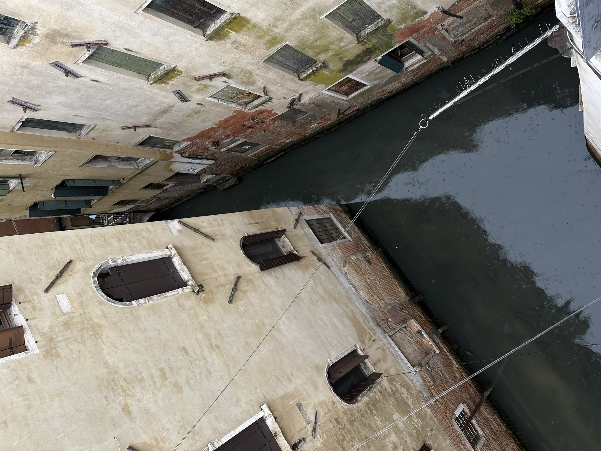 The image shows a narrow canal in Venice, Italy, flanked by tall buildings. The buildings are old and weathered, with a variety of windows, some boarded up or shuttered. The water in the canal is dark and reflects the sky. A line is strung across the canal, possibly for mooring boats. There are no people visible in the image. The perspective is looking down into the canal from above.
