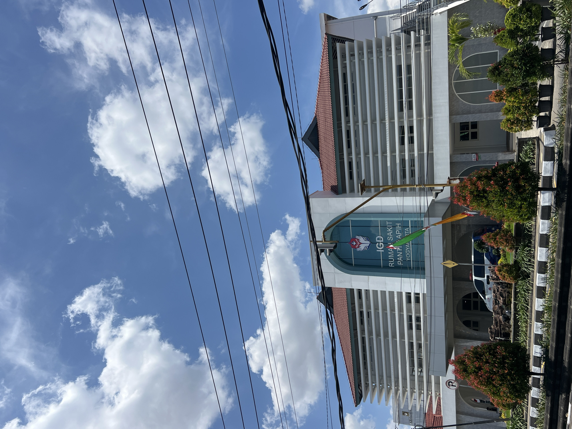 The image captures the exterior of the IGD (Instalasi Gawat Darurat - Emergency Room) of Rumah Sakit Panti Rapih in Yogyakarta, Indonesia. The building has a modern architectural style with white louvers on its facade and a prominent red-tiled roof. The entrance area features a large arched window with the hospital's logo and text that reads "IGD RUMAH SAKIT PANTI RAPIH YOGYAKARTA".
In front of the building, a line of well-maintained trees are planted in pots, creating a neat landscaping. An ambulance is visible parked near the entrance, indicating its operational status. The sky above is a vibrant blue, dotted with fluffy white clouds. Power lines are visible in the foreground, stretching across the frame and slightly obscuring the view of the building and sky. The overall scene suggests a bright, clear day in an urban setting.