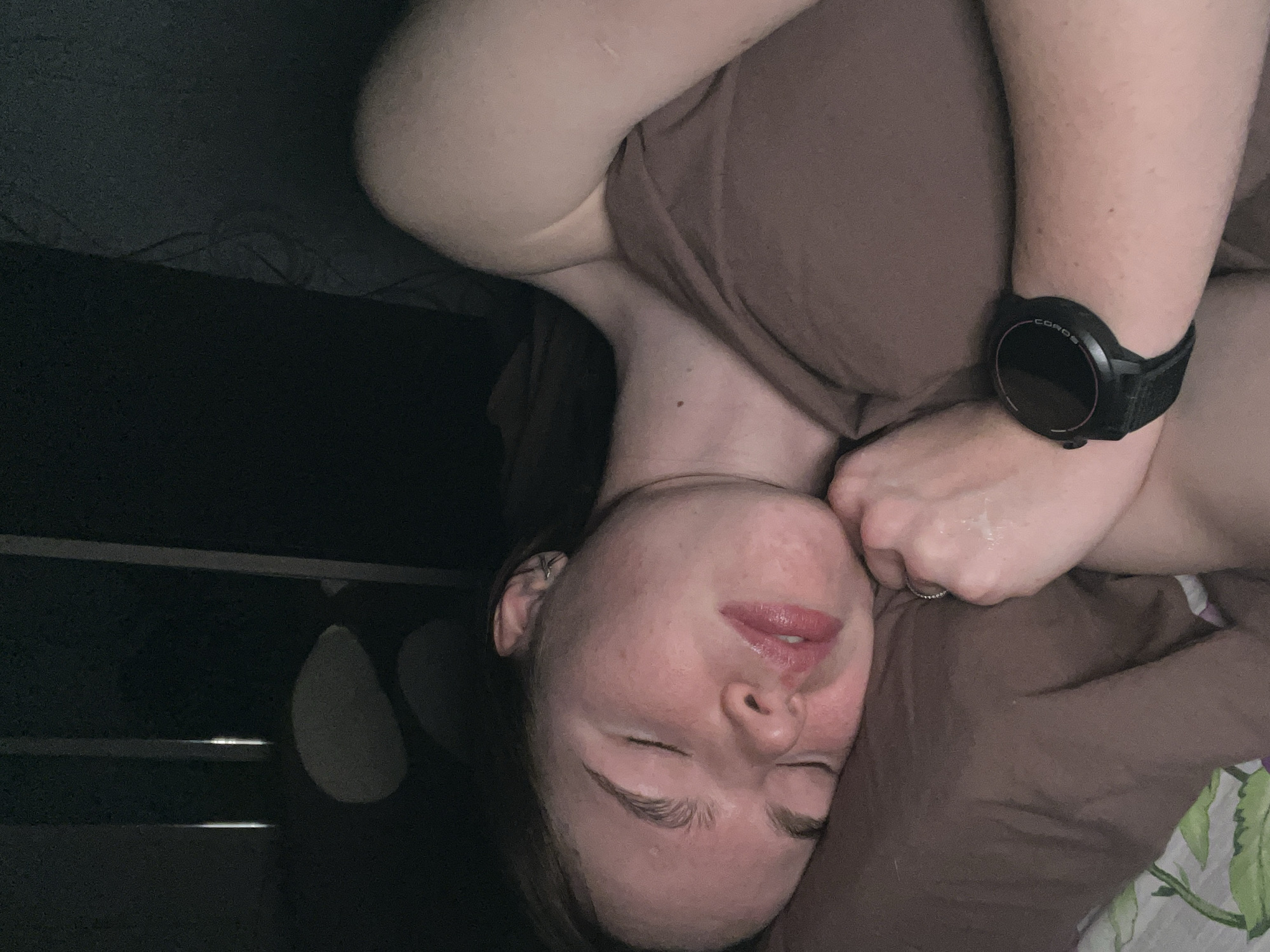 An individual, identified as likely female, is depicted lying on their right side with closed eyes, indicating sleep or rest, in a dimly lit indoor environment within the city of Chaikovsky, Russia. The person exhibits fair skin with minor facial redness. Dark hair frames the head, and a silver-colored piercing or ear cuff is visible on the helix of the right ear. The lips are slightly parted. The subject is wearing a muted reddish-brown garment. A mole is present on the right side of the neck/shoulder area. The left arm is bent, with a silver-colored ring worn on a finger of the left hand. A black COROS brand smartwatch with a black strap is visible on the left wrist. The individual rests on a brown pillow, which in turn lies on a white sheet adorned with a green floral pattern. The background is dark and indistinct, featuring several faint vertical illuminated strips in the upper left quadrant.