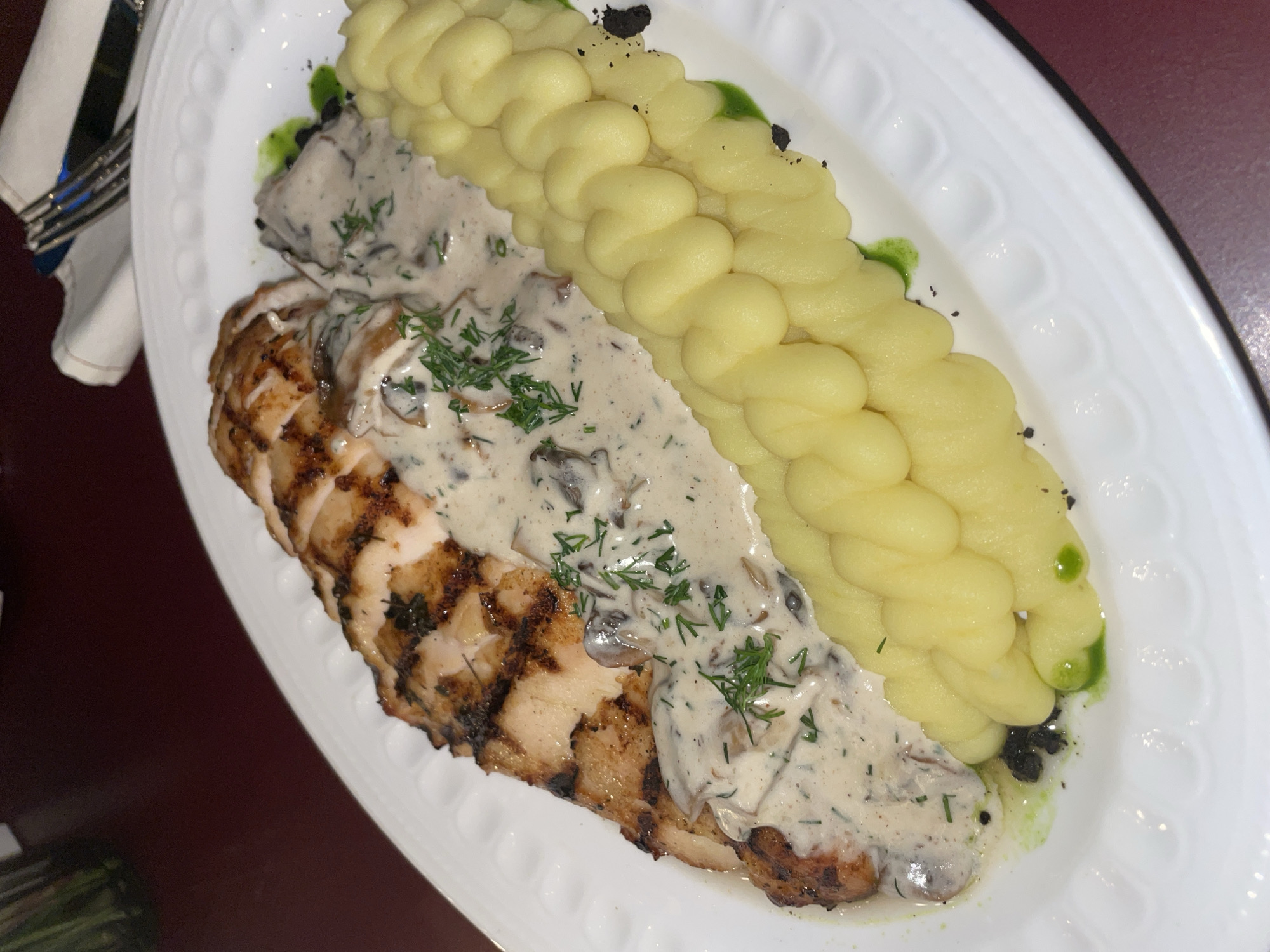 The image presents a plated dish featuring grilled chicken breast, topped with a creamy mushroom sauce, and accompanied by mashed potatoes, likely served in a restaurant setting. The chicken displays distinct grill marks, indicating a cooking process that produced a slightly charred exterior. The mushroom sauce is generously applied, suggesting a rich and savory flavor profile. The mashed potatoes are piped into a decorative design. The dish is served on a white, oval-shaped plate with an embossed rim. Based on the provided information, it is impossible to definitively confirm that the image was taken in Yerevan, Armenia. The location cannot be verified from the visual information alone.