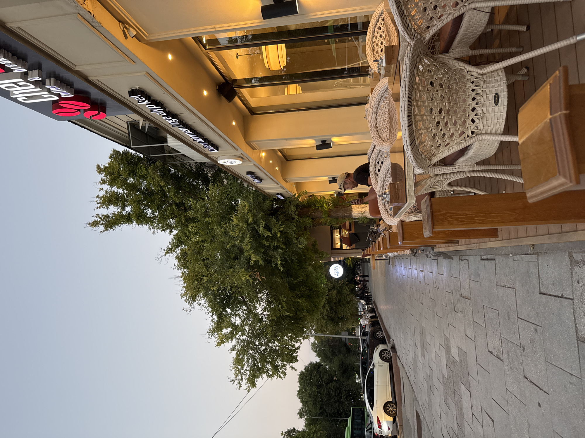 The image depicts an outdoor seating area of a coffee shop, likely located in Shymkent, Kazakhstan, given the signage. The area features several tables and chairs made of woven material, with some occupied by patrons. One man is visible sitting at a table, looking at his phone. The coffee shop itself has a modern facade with large windows and illuminated signage. In the background, a sidewalk lined with trees separates the coffee shop from a street with parked cars and passing traffic. The sky is clear and the overall atmosphere suggests a late afternoon or early evening setting.