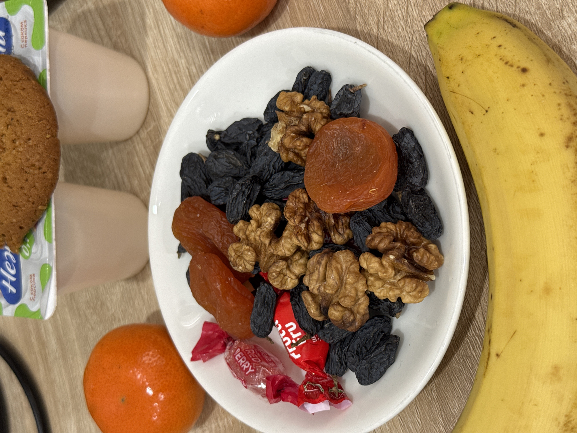 This is a top-down view of a table laden with food, specifically a light brown surface. A white dish dominates the central area, filled with a mixture of dark raisins, walnuts, and dried apricots. Two wrapped red candies, labeled "RED BERRY" and "frutto," are positioned on the bottom edge of the dish. To the right of the dish is a yellow banana. To the left, a cookie is placed next to two small containers. Two mandarin oranges are also visible. Based on the branding of the item next to the cookie, which features Cyrillic text, it's likely this photo was taken in a location that uses the Russian language. As such, this scene could be from Tashkent, Uzbekistan.
