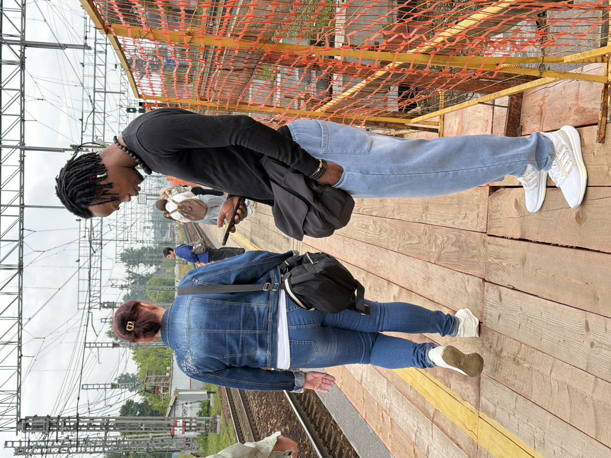 At a railway platform in Shcherbinka, Russia, a Black man with dreadlocks, wearing a black long-sleeved shirt and light blue sweatpants, stands near an orange safety net. He is looking down at his phone and has a black bag slung over his shoulder. To his left, a woman with red hair, wearing a denim jacket and blue jeans, walks away from the camera. She also carries a black bag. The platform is made of wooden planks with a yellow line marking the edge. Overhead, numerous electrical wires and support structures are visible, indicative of a train station environment. Other people are visible in the background, some standing and others walking. The sky is overcast.