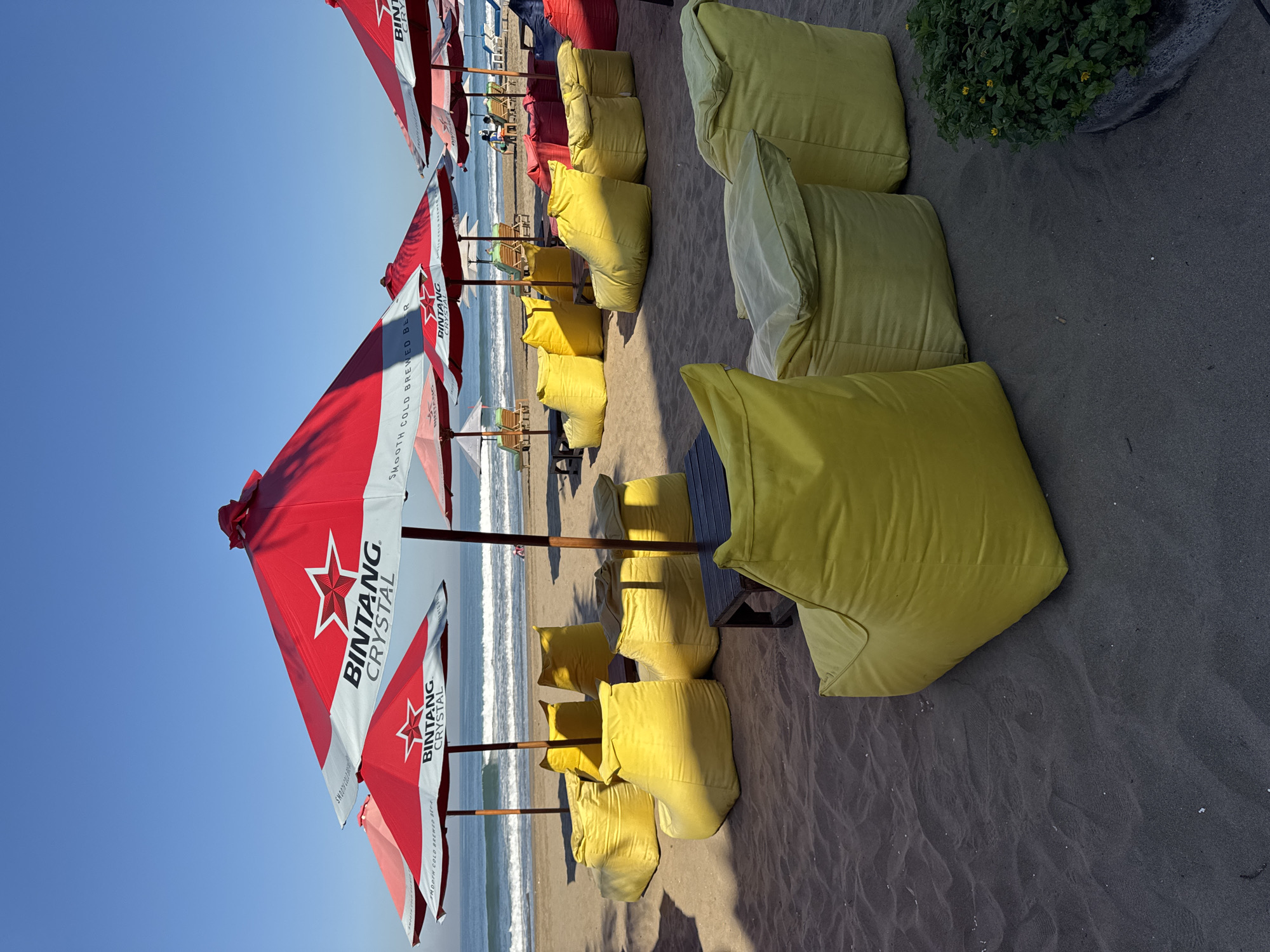 The image shows a beach scene in Kuta, Indonesia, with numerous red and white Bintang beer umbrellas and yellow beanbag chairs lined up on the sand. The beach is characterized by dark sand and gentle waves. In the background, there are additional umbrellas, sun loungers, and a few people visible on the beach. The sky is clear and blue, suggesting a sunny day. The arrangement of the umbrellas and beanbags indicates a designated seating area.