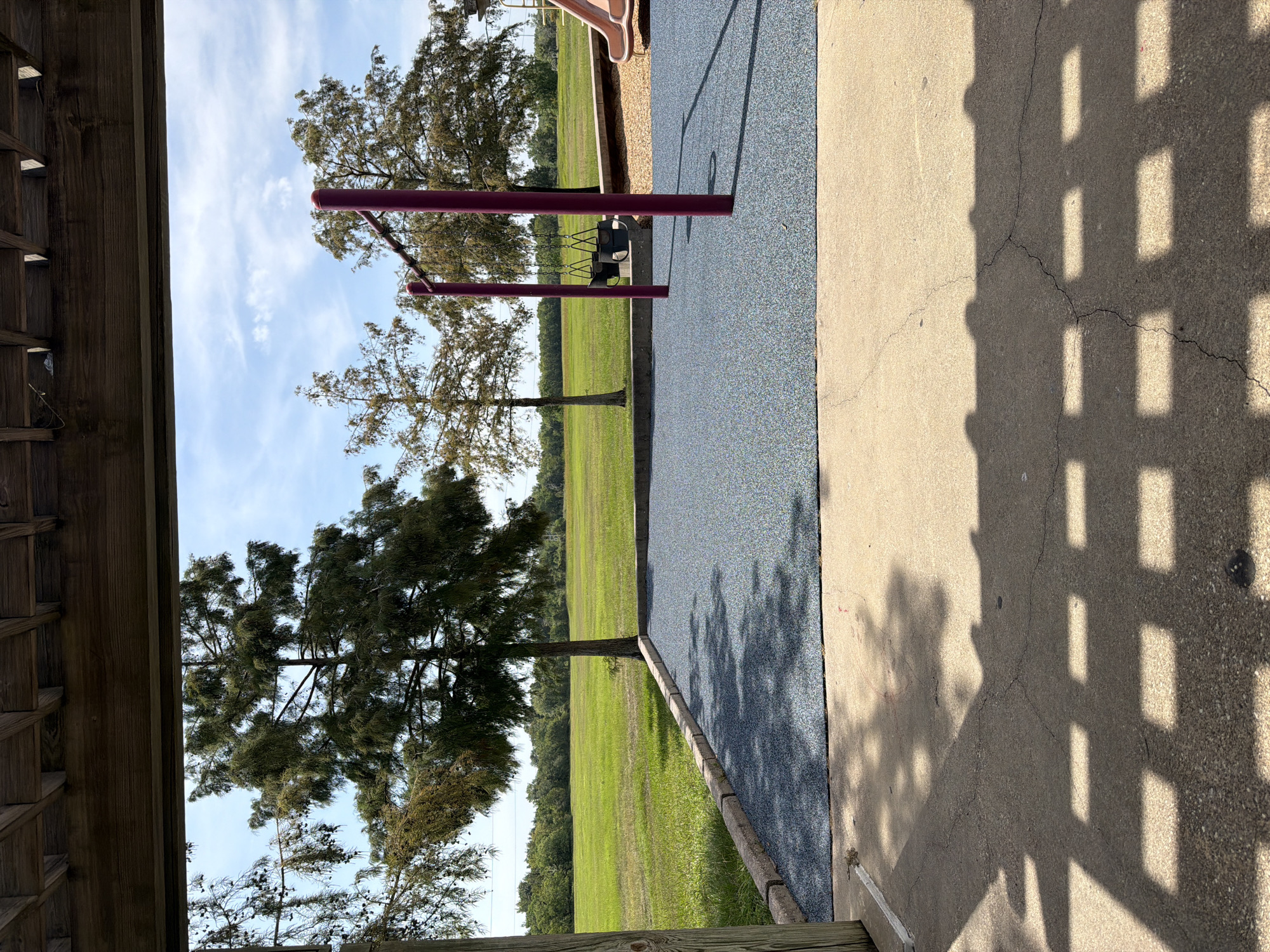 The image depicts an outdoor scene at a playground. In the foreground, a concrete patio with a shadow pattern cast by an overhead structure is visible. Beyond the patio is a playground surface made of blue rubberized material, bordered by concrete curbing. A set of purple swing set poles with two black swings hanging from them stands on the blue surface. To the right, a portion of a pink slide is visible. In the midground, several trees are present, with a large expanse of green grass stretching into the background. The sky above is blue with scattered white clouds. The overall impression is that of a sunny day at a park. The location is a playground in Houston, Texas, United States of America.