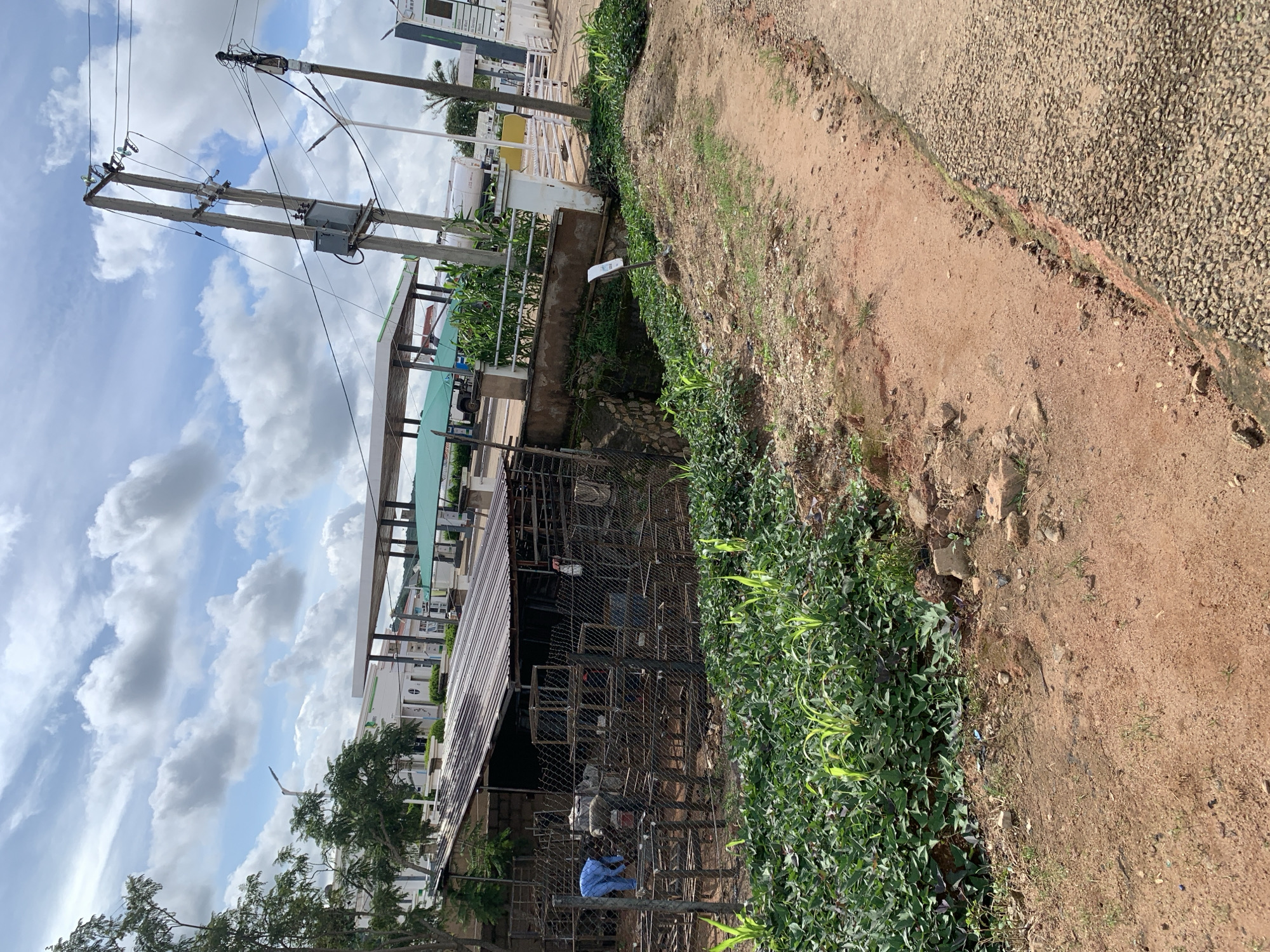 The image depicts an outdoor scene, possibly on the outskirts of a city. In the foreground, a dirt path with scattered rocks and debris runs alongside a paved road. To the left, a densely vegetated area with green plants and some flowering vines is visible.
Further back, a rustic, open-sided structure with a corrugated metal roof is present. This structure appears to be a workshop or storage area, with metal frames and wire mesh enclosures visible. A person wearing a blue garment is working within this structure, near some metalwork.
Behind the workshop, a gas station with covered pumps and a white building is discernible. Power lines and utility poles with transformers are prominent in the background. The sky is partly cloudy with scattered white clouds against a blue backdrop. The overall impression is one of a functional, somewhat informal area, possibly undergoing construction or repair.