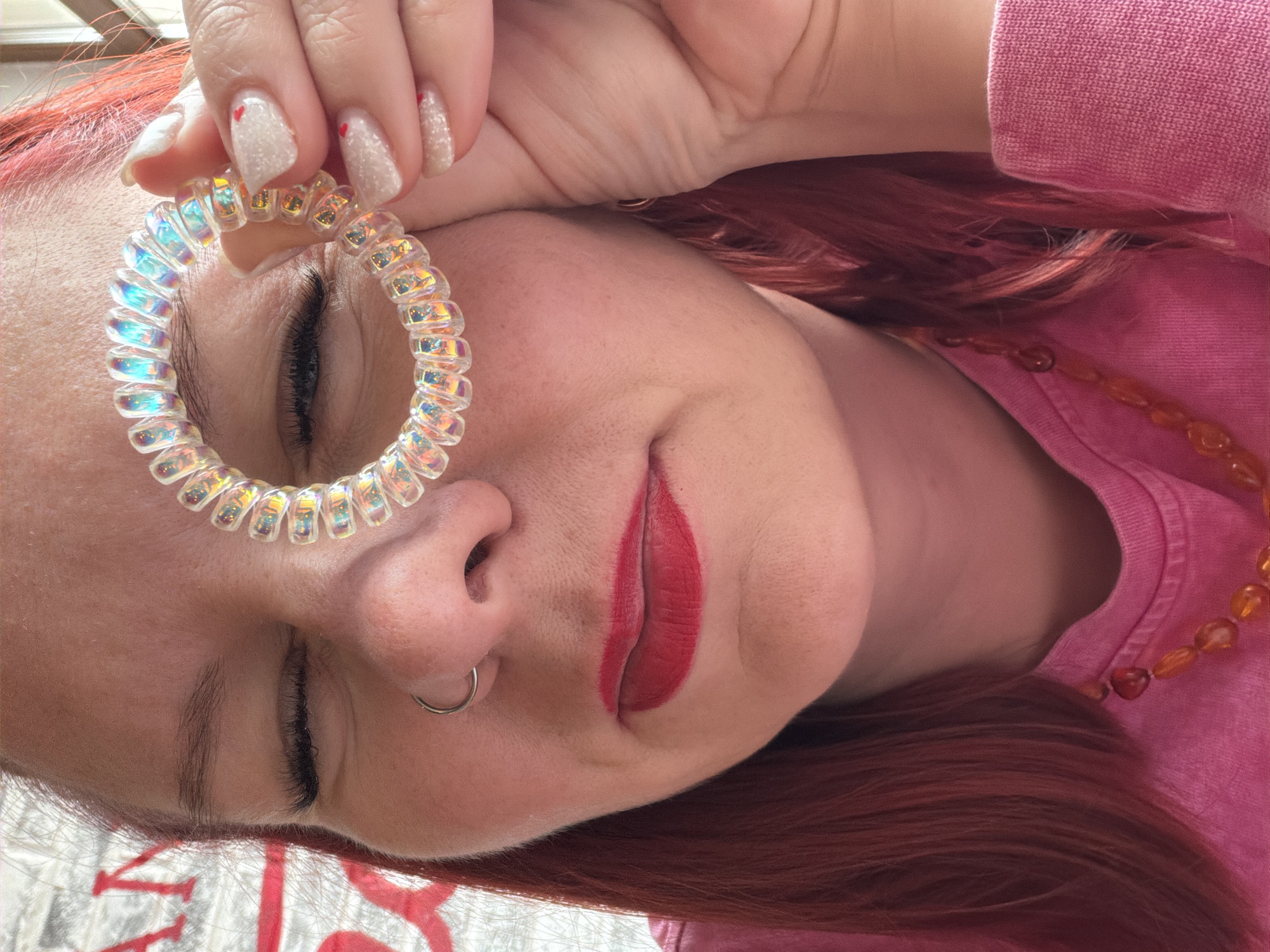 A woman with reddish hair and fair skin is lying down. Her eyes are closed. She is wearing red lipstick and has a silver hoop piercing in her left nostril. She is holding a transparent, iridescent spiral hair tie over her left eye. Her nails are decorated with glitter. She is wearing a pink shirt and a necklace with orange beads. The image may have been taken indoors. There is no information regarding the location being in Chaikovsky, Russia.