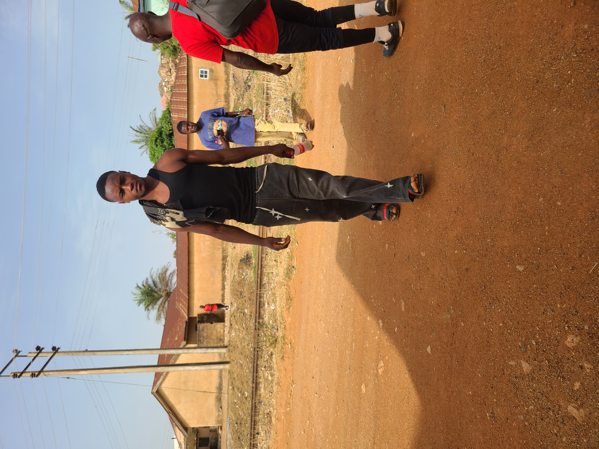 The image shows three men walking along a dirt road in Jos, Nigeria. The man closest to the camera is wearing a black tank top and dark jeans with star patterns. He is holding a red bottle in his right hand. Behind him, a second man is wearing a red shirt and black pants, carrying a black backpack. Further back is a third man in a blue shirt and khaki pants, holding a smartphone. The road is lined with buildings and vegetation. Power lines and poles are visible on the left side of the image. A set of railroad tracks runs between the road and the buildings. The sky is clear and blue.