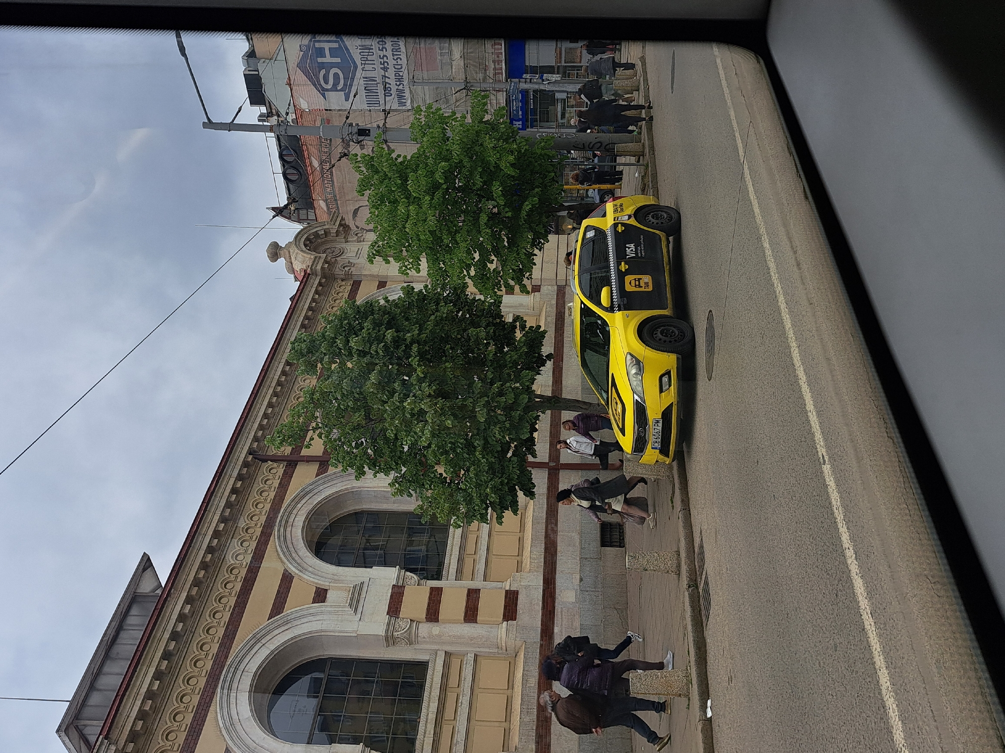 Here's a detailed description of the image:
The photo, likely taken from inside a vehicle, presents a street scene in Sofia, Bulgaria. A yellow taxi cab is parked along the curb, partially obscured by two trees. The cab has "VISA" and "TAXI ME" written on its side, and its license plate reads "CB 6067 PM". Several pedestrians are walking on the sidewalk near the taxi, some passing behind the trees and others closer to the building.
The building alongside the sidewalk is an older structure with large arched windows. Its facade is adorned with decorative architectural details in beige, cream, and red/brown. A sign with the text "SH ШПИЦИ СТРОЙ 0877-455 505 WWW.SHPICI-STROI." is visible on a building across the street, indicating a construction-related business. The sky is overcast, with a pale grey color. The image is framed by the window or interior structure of the car from which it was taken.