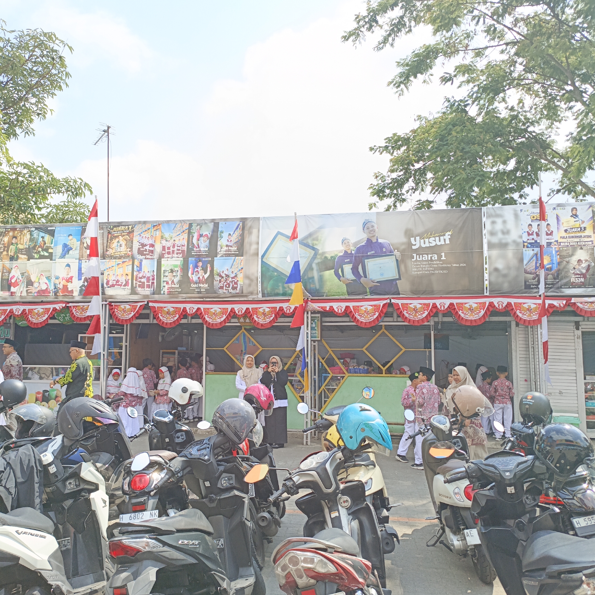 The image depicts an outdoor scene in front of a building adorned with banners and decorative red and white pennants, suggestive of a public event or celebration, likely in Salatiga, Indonesia. The banners feature images of people, awards, and text, indicating a possible school or community event. In the foreground, numerous motorcycles are parked, some with helmets placed on them. Several individuals, including children and adults dressed in traditional attire or uniforms, are visible interacting near the building. The atmosphere appears to be daytime with clear skies, with trees framing the top of the image. The overall impression is of a vibrant gathering, possibly a school award ceremony or festival, taking place in a public area in Salatiga.