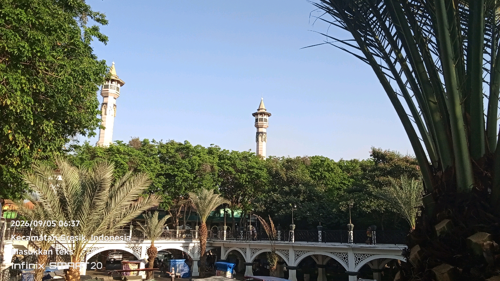This image depicts a sunny outdoor scene in Gresik, Indonesia, on September 5, 2026, at 06:37. In the background, two minarets of a mosque rise above lush green trees. A decorative white bridge with arched supports spans across a body of water, with people visible walking on it. Palm trees are prominent in the foreground, with one large palm tree obscuring a portion of the right side of the frame. The overall environment suggests a park or public area adjacent to the mosque. The text overlay indicates "Kecamatan Gresik, Indonesia" and "Infinix Smart 20."