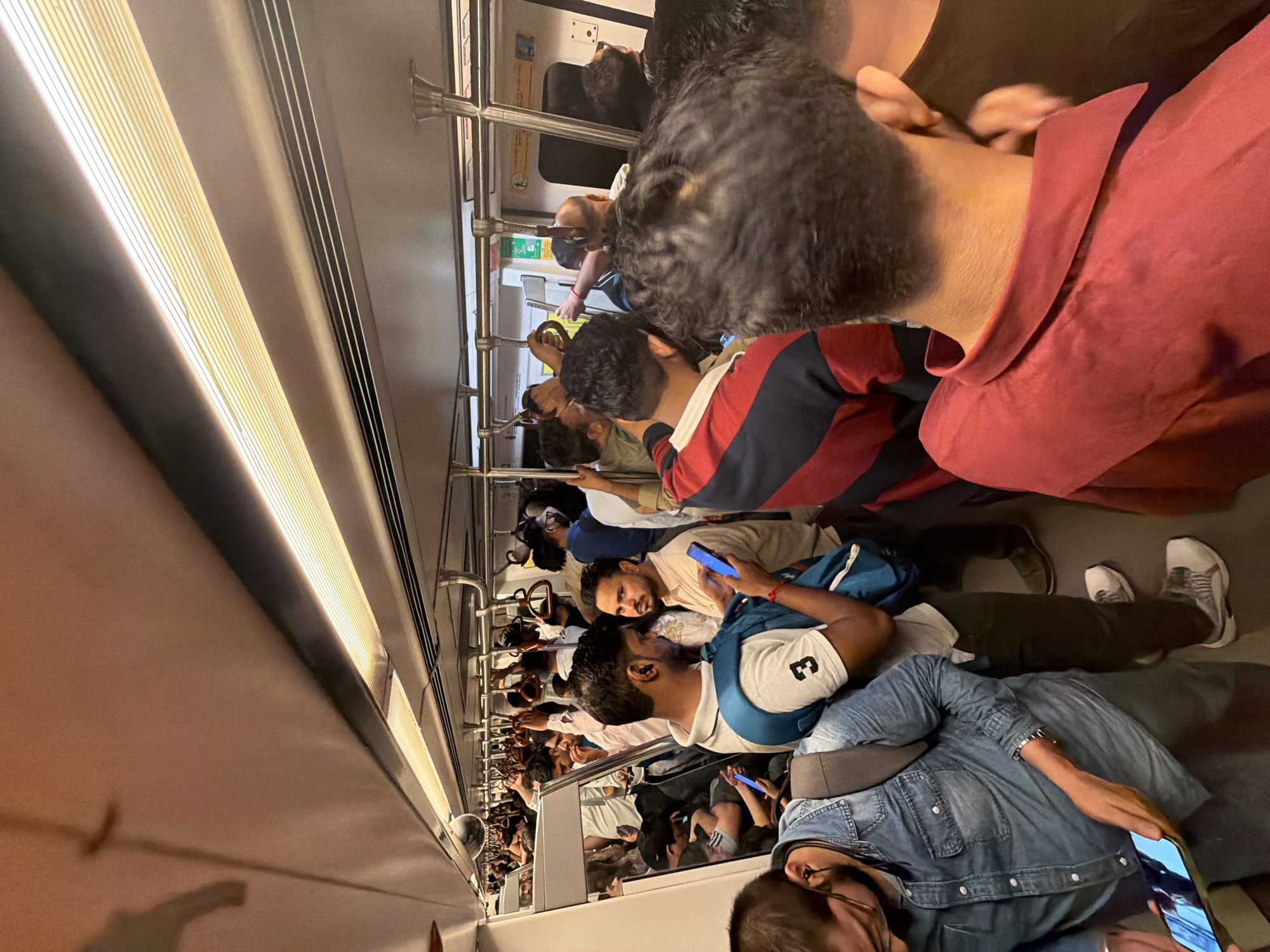 This is a photograph taken inside a crowded Delhi Metro train. The train car is filled with passengers, many of whom are standing and holding onto the overhead handrails. People are dressed in casual clothing, and some are looking at their mobile phones. The environment is typical of a public transit setting, with fluorescent lighting and metal fixtures. The image captures a moment of daily commute in the bustling city of Delhi, India, highlighting the dense population and the widespread use of public transportation.