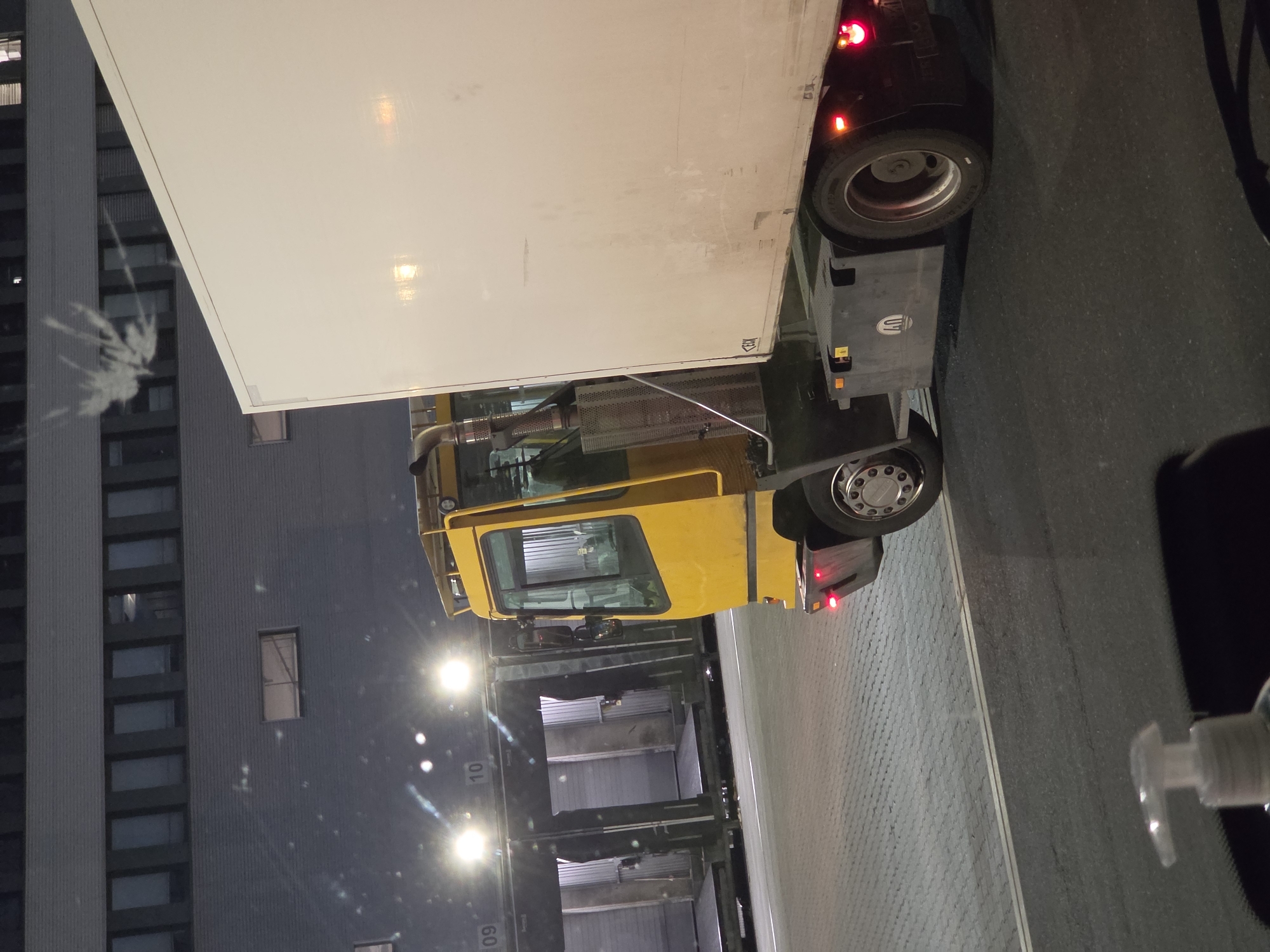 The image shows a yellow truck cab with a white trailer parked in front of loading docks at night. The loading docks are numbered 09 and 10. The truck is positioned to the right of the loading docks. The background shows a modern building with many windows. The ground is paved with grey bricks. A hand sanitizer bottle is visible in the foreground, suggesting the photo was taken from inside a vehicle. The scene appears to be a logistics or distribution center.