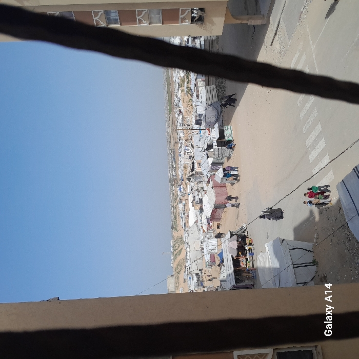 The image displays an elevated view of a dense, temporary settlement or refugee camp in Khan Yunis, Palestinian Territory, under a clear, pale blue sky. The scene is framed vertically by a light brown building column on the left and a darker brown, narrower building column on the right, both suggestive of a window or balcony enclosure. A thin black cable extends diagonally from the upper left to the lower right across the central view. The "Galaxy A14" watermark is visible in the lower-left corner.
In the foreground, a wide dirt road is present, featuring a faded white pedestrian crossing marked with parallel lines. A group of three to four children stands on the dirt road near the pedestrian crossing. Further along the road, an adult figure in dark clothing walks from left to right, casting a shadow. To the immediate left of the road, a makeshift stall with a white tarp roof and various goods displayed underneath is visible. To the right of the pedestrian crossing, a dark horse, possibly with a person, is located on the dirt path.
The midground is densely packed with hundreds of makeshift shelters, shanties, and tents constructed from a variety of materials including corrugated metal, tarpaulins, and fabric, extending across a vast, sandy, and arid landscape. Individuals are observed walking amidst these structures. Some structures appear to be market stalls with canopies. On the far right, a portion of a multi-story, light brown building with multiple windows, some appearing damaged or boarded, is visible.
In the background, the temporary settlement continues towards a flat horizon. Beyond the camp, the terrain is characterized by lighter-colored, dry ground with sparse vegetation or distant structures, and subtle elevated mounds are discernible against the sky.