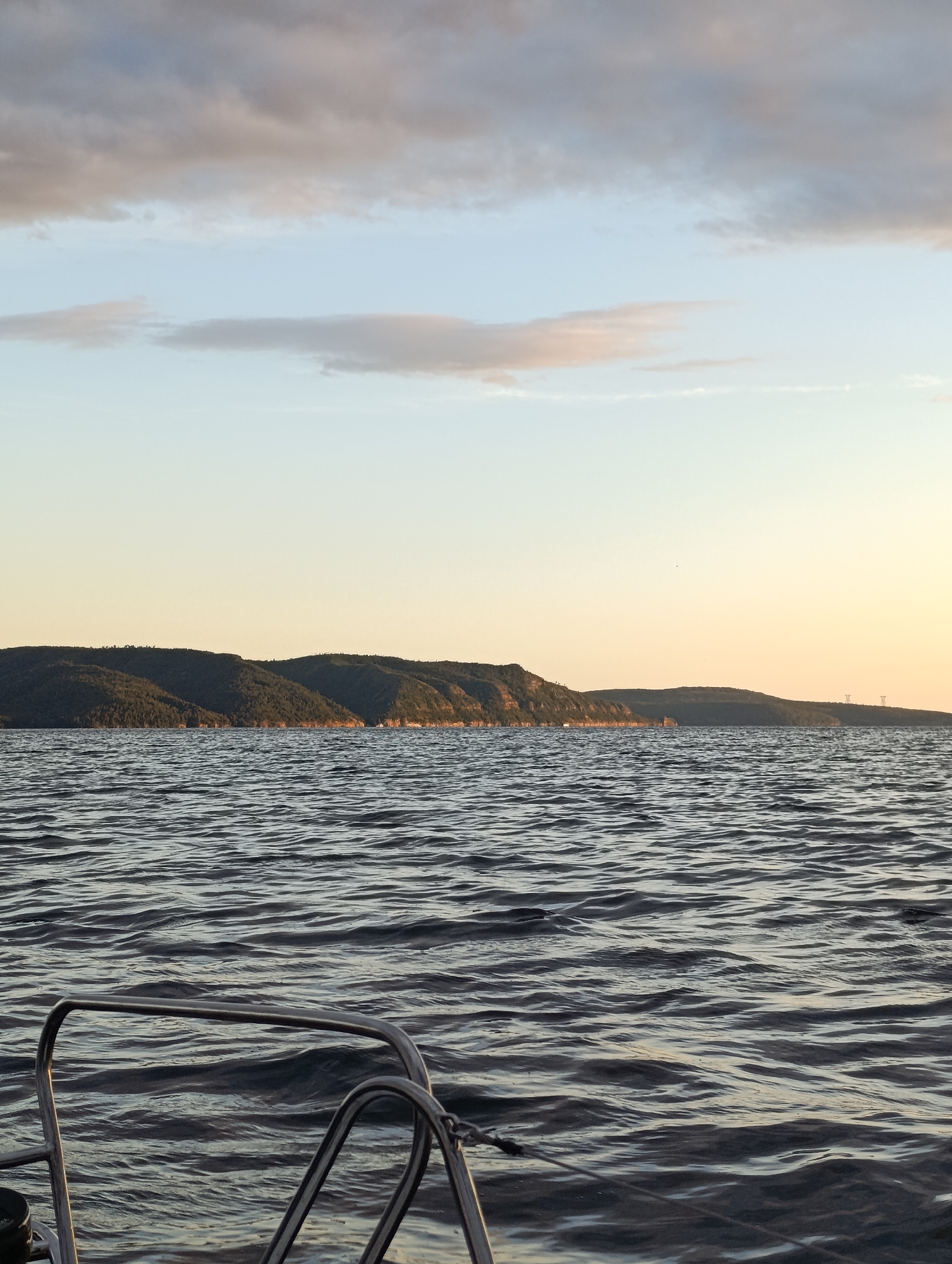 The image shows a view from a boat on the Volga River near Tolyatti, Russia. In the foreground, metal railings of the boat are visible, along with the rippling water of the river. In the middle ground, a long, forested embankment with undulating hills stretches across the horizon. The sky above is a pale blue with scattered clouds, transitioning to a soft yellow hue near the horizon, indicating either sunrise or sunset. The overall scene depicts a tranquil natural landscape viewed from the water.