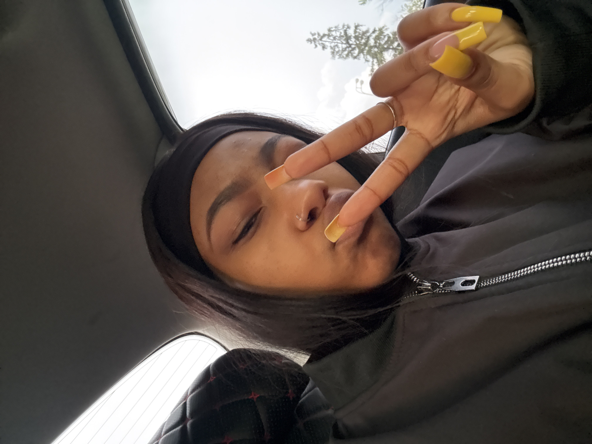 A young woman poses for a selfie inside a vehicle. She wears a black headband and a black jacket with a zipper. Her long, dark hair frames her face. She holds up her hand in a peace sign, displaying yellow-tipped, square-shaped nails, and wears a silver ring. Her eyes are partially closed, and her lips are pursed in a playful expression. The vehicle interior consists of a dark headliner and a seat with black quilted upholstery featuring red stitching. The background suggests an outdoor environment with a bright sky and possibly trees visible through the vehicle's roof. There is no information to place the location in the city of Troitsk, Russia.