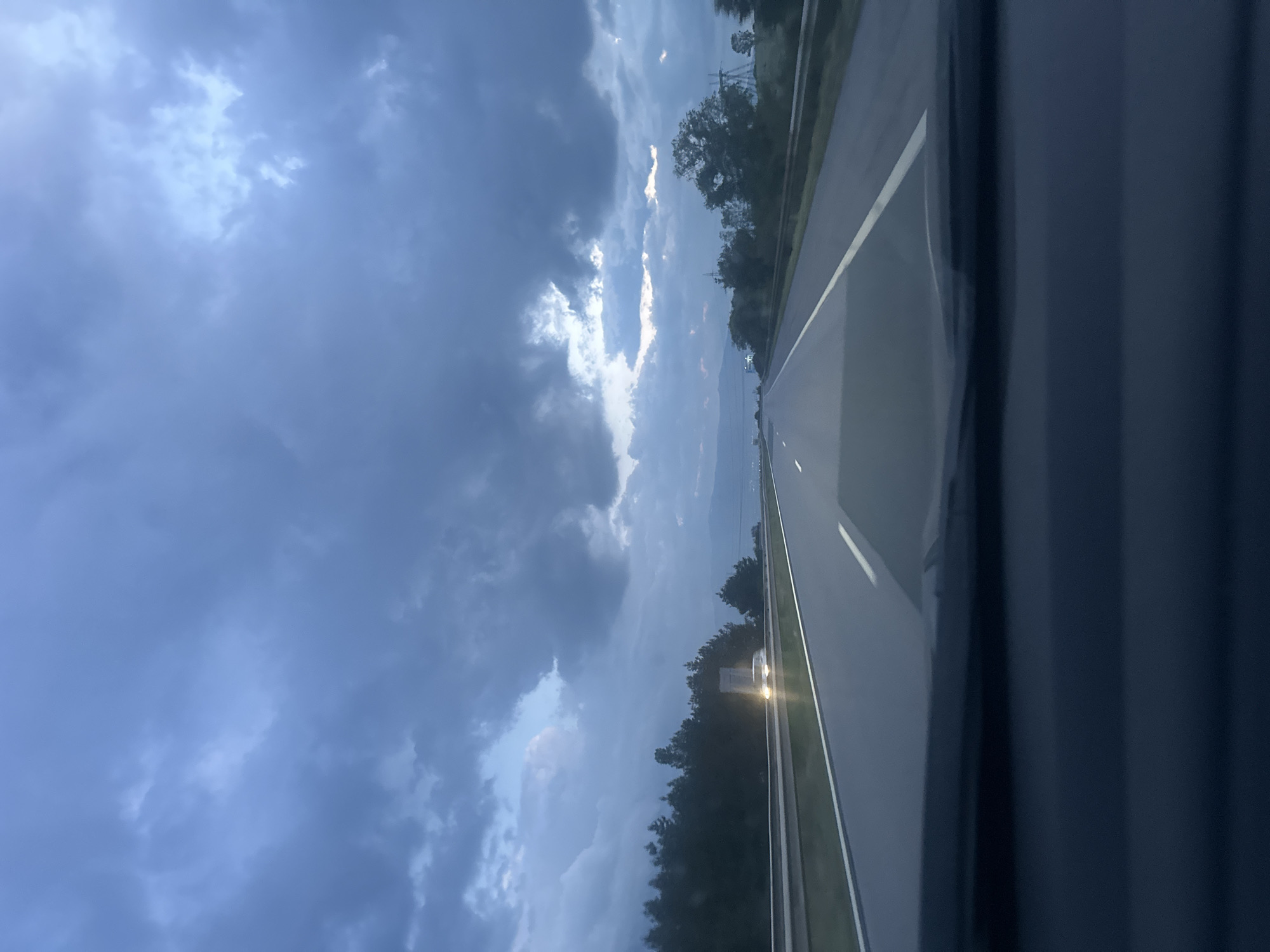 The image presents a view from inside a vehicle, looking out onto a highway, with the photograph appearing to be rotated 90 degrees counter-clockwise.
The sky dominates the upper and left portions of the frame, filled with dense, dark gray clouds that suggest an overcast or stormy weather system. However, brighter patches of sky and sunlit clouds are visible breaking through the gloom, particularly towards the upper left and mid-left, indicating that some light is penetrating or that a break in the cloud cover exists.
The highway occupies the lower and right section of the image, stretching into the distance. It features clear asphalt and distinct white road markings, including a solid line on the immediate right shoulder and a broken line further to the left, separating lanes. A metal guard rail is visible along the right side of the road. Both sides of the highway are flanked by dense green trees and foliage, suggesting a rural or forested landscape. In the far distance, along the right side of the road, the faint glow of headlights from an approaching or distant vehicle can be seen. A dark, vertical pillar, likely part of the vehicle's interior frame, is visible along the far right edge of the image.
The time of day appears to be late evening or dusk, evidenced by the muted light, the nature of the heavy clouds, and the visible vehicle headlights. The weather is heavily clouded but with glimmers of brighter light. The scene depicts travel on a highway, potentially in the area of Zhelyava, Bulgaria, as provided by the context. There is no visible text in the image.