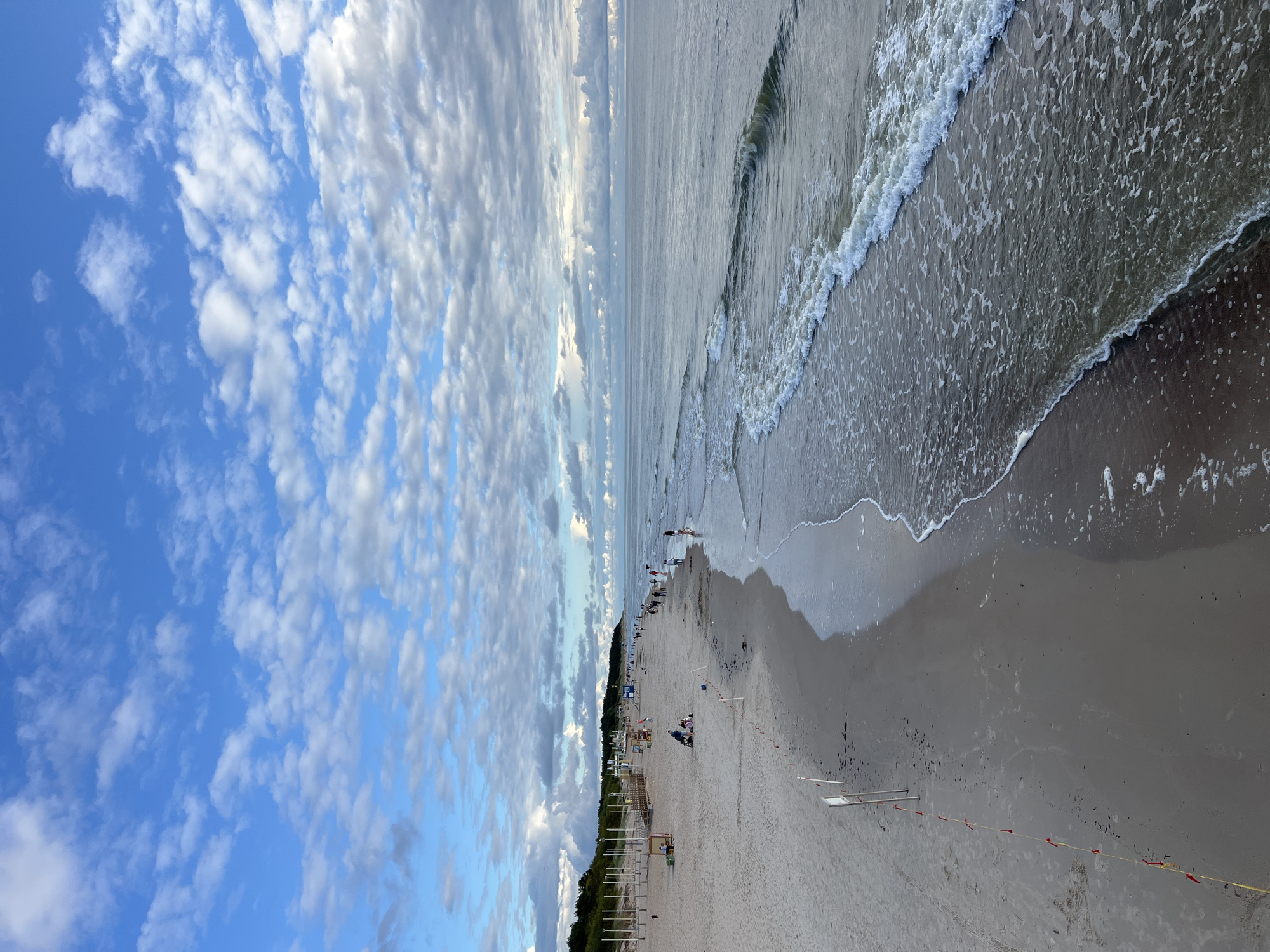 The image captures a wide, sandy beach in Palanga, Lithuania, under a sky filled with scattered clouds against a bright blue background. The sea is calm with gentle waves lapping at the shore, creating a foamy white line as they recede. The beach is sparsely populated with a few individuals and families scattered across the sand. Some people are walking along the water's edge, while others are seated or standing further up on the beach.
In the background, a line of trees forms a natural border separating the beach from a resort area. There are structures that appear to be beach amenities, such as wooden walkways or piers, and some small shelters. Red and yellow flags on poles mark a boundary on the sand, possibly indicating a restricted area or hazard. The overall impression is one of a peaceful, late afternoon scene at a seaside resort.