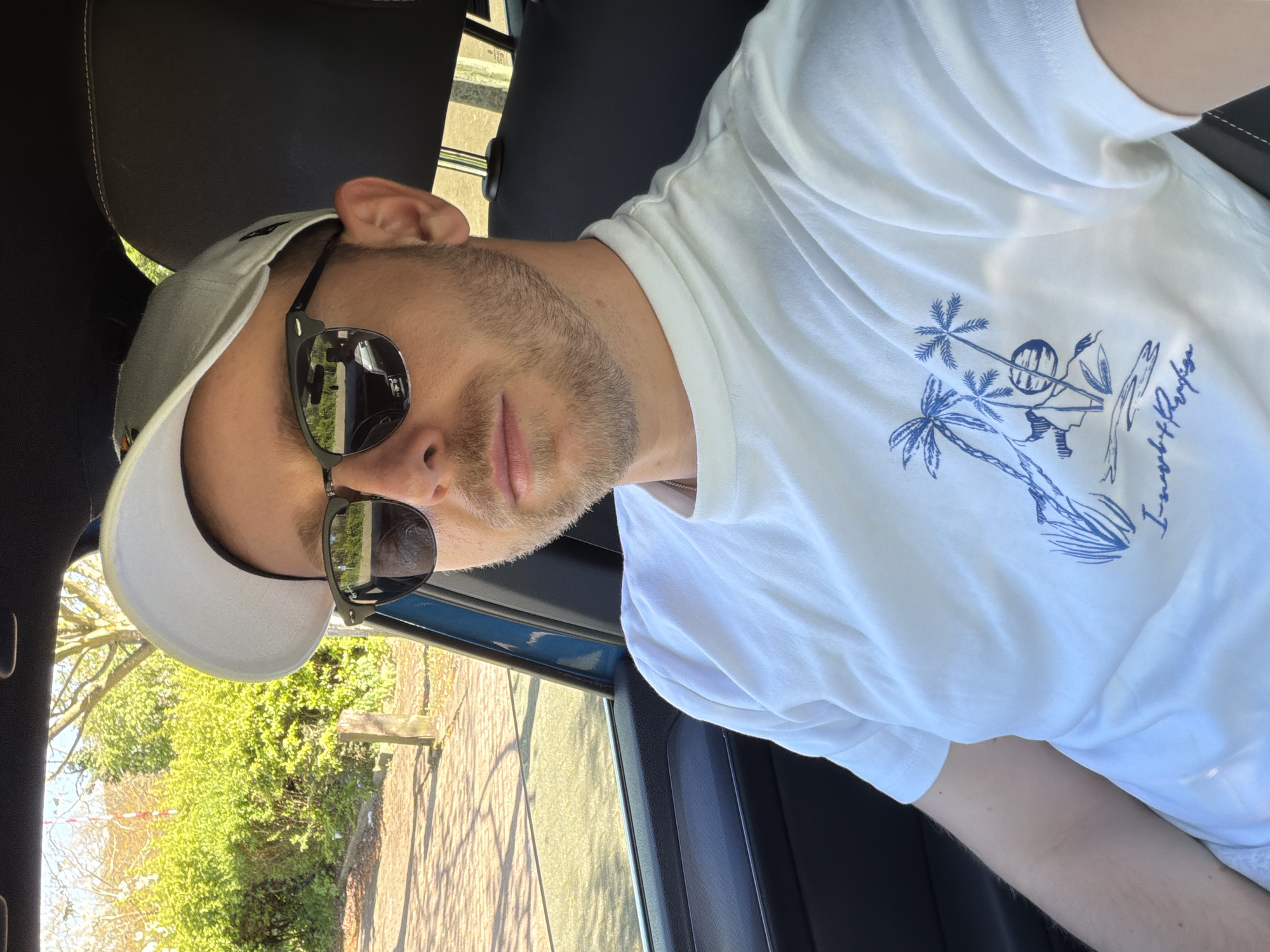 A man is seated in a car. He wears a white t-shirt with a blue tropical-themed graphic, a light-colored baseball cap, and sunglasses. He has short, light brown hair and stubble. The car is parked on a street; trees and shrubs are visible through the open car window. It is daytime, and the lighting is bright. There is no evidence in the image to indicate that the location is London, United Kingdom.