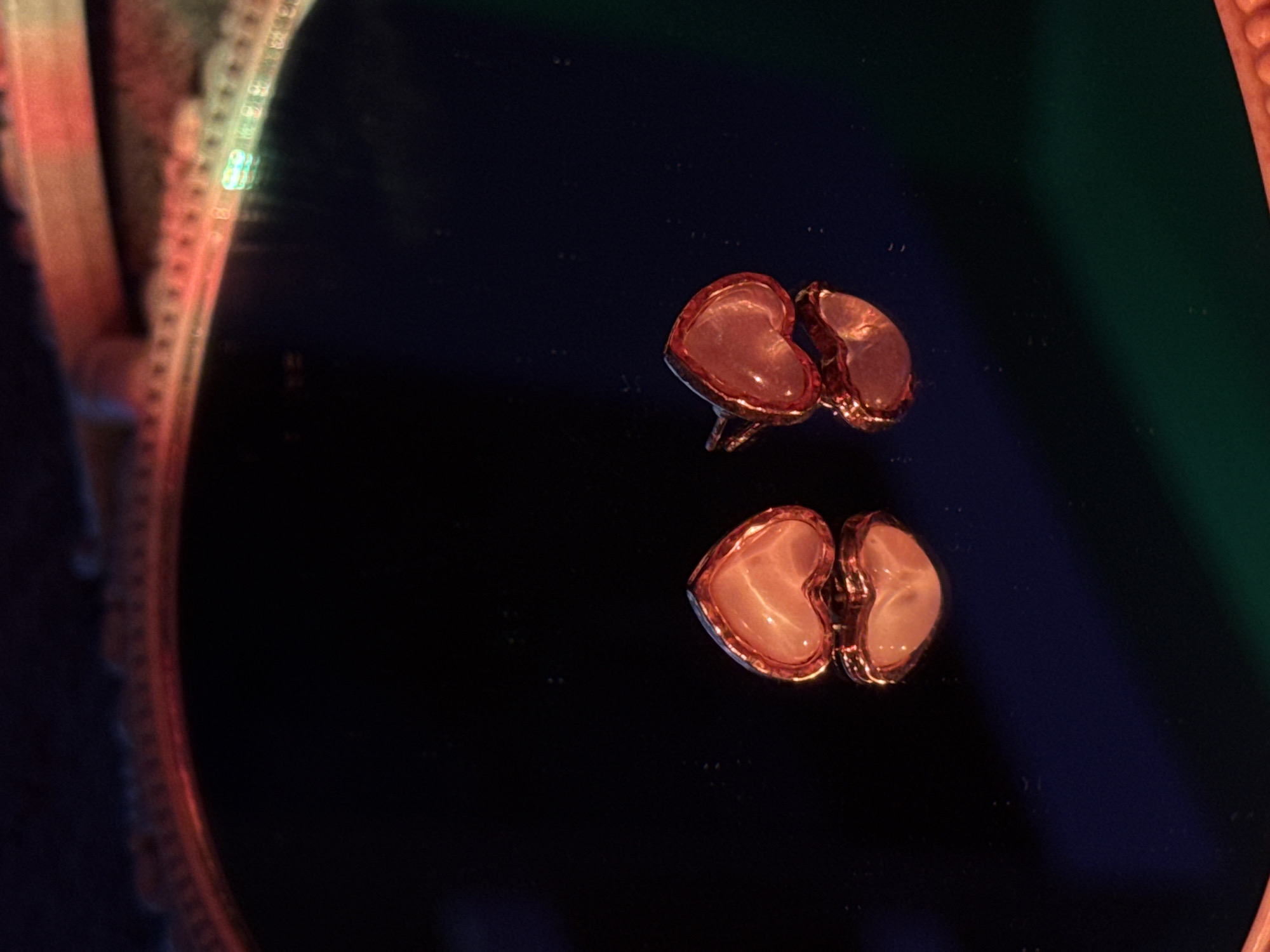 The image shows a pair of heart-shaped earrings with a pinkish-white stone and a gold-colored frame. They are reflected in a dark, glossy surface, likely a mirror. The edge of the mirror frame is visible on the left side of the image, and a green surface is seen in the upper right. The location in the city is unknown.
