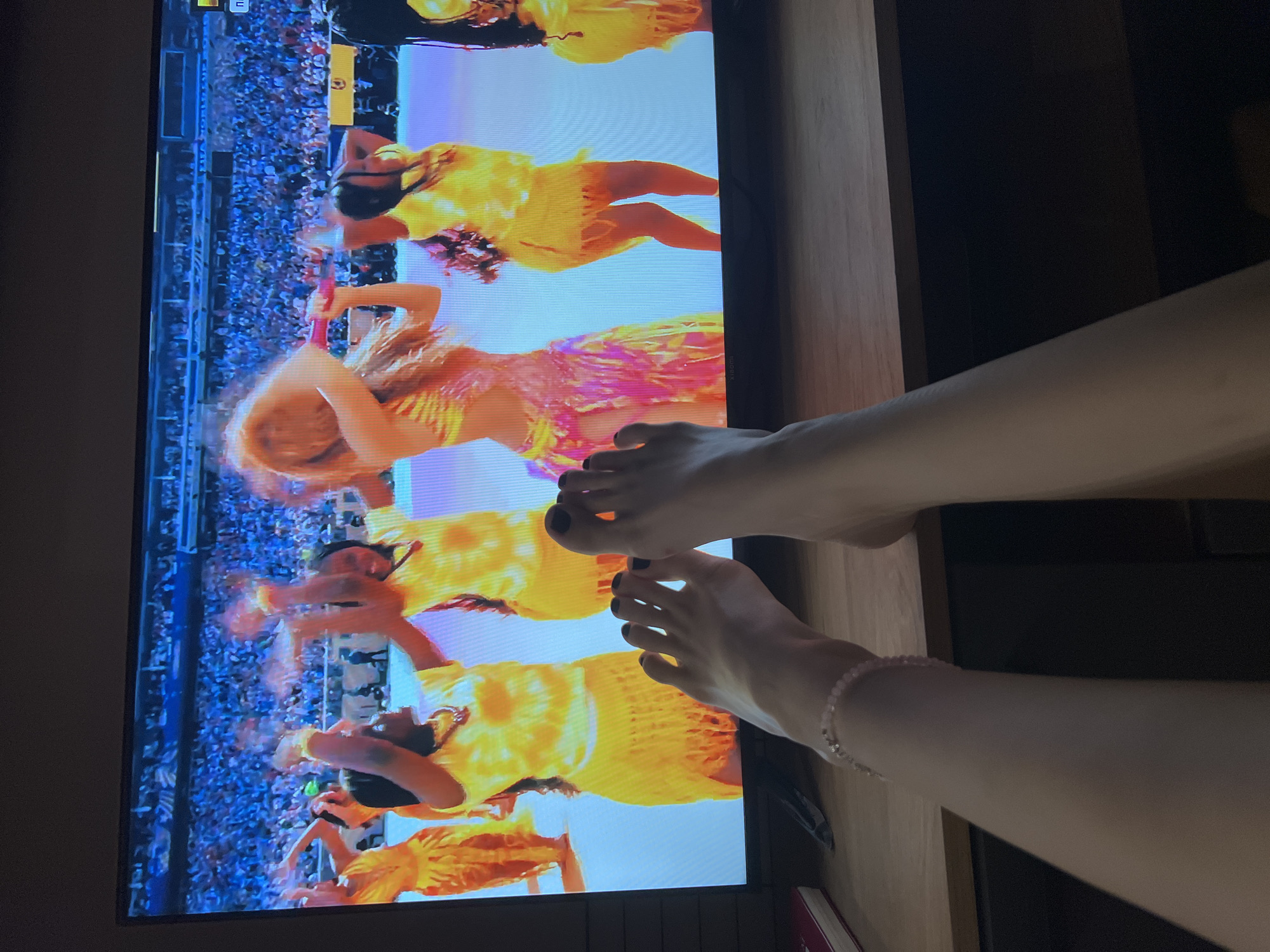 A first-person perspective shot shows a pair of bare feet with black nail polish resting on a wooden surface in the foreground, with a television screen displaying a vibrant performance in the background. The performance features several women in bright yellow and pink outfits, with one woman in the center with long, flowing reddish-brown hair. They are on what appears to be a stage or stadium with a large crowd visible in the upper portion of the screen. The television is placed on a desk or table that is made of wood, and a book with a red cover is visible on the left side of the desk. The lighting suggests an indoor setting, likely a living room or entertainment area. There is no indication of the location being in the city of Moscow, Russia.