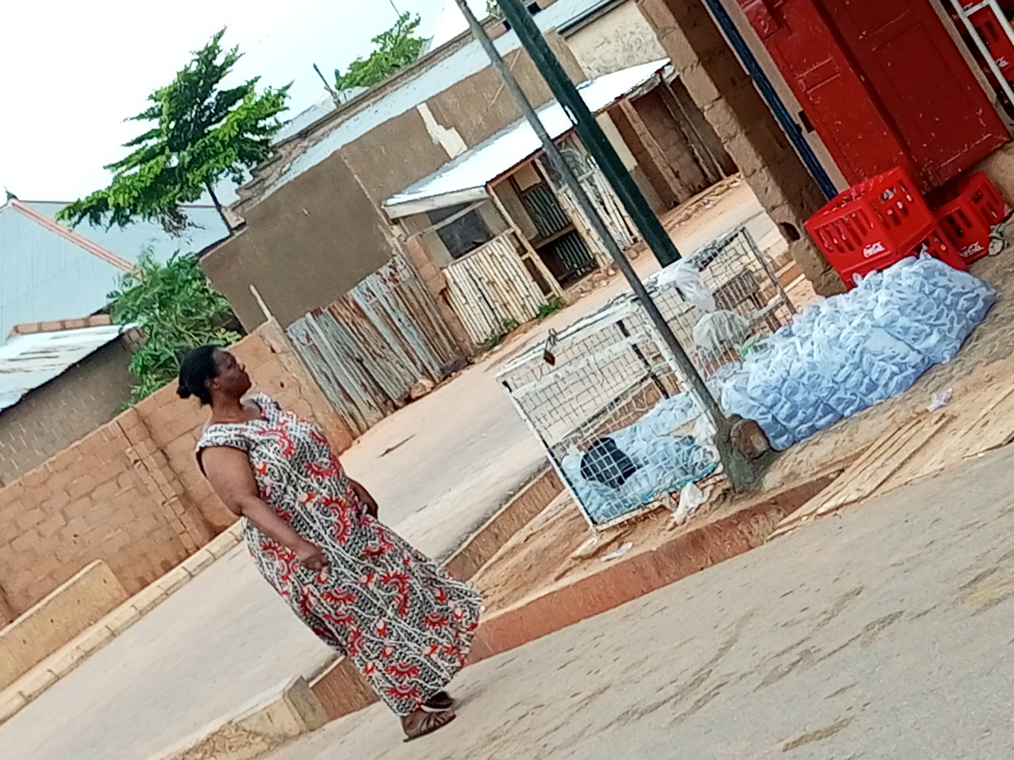 A woman in a patterned dress walks across a dusty, uneven street in Tempure, Nigeria. She is angled away from the camera, looking to her right. To her right, on the side of the street, are stacked red Coca-Cola crates and a large quantity of bagged white material, possibly water sachets. Behind the bagged material is a metal cage containing a person. In the background are buildings with corrugated metal roofs and brick walls. A small tree with green leaves grows from a rooftop. The sky is overcast.