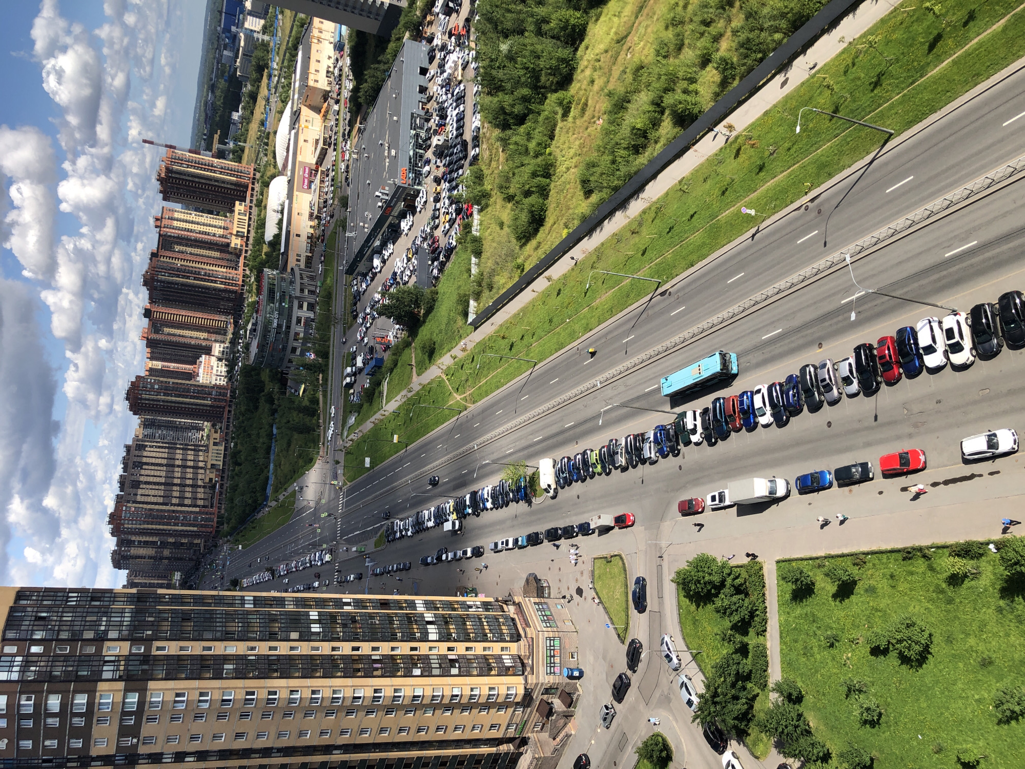 The image is an aerial view of a multi-lane road in an urban environment. A tall, modern residential building with a beige and brown facade dominates the left side of the frame. On the right, a long row of parked cars lines the roadside, followed by a grassy verge and a dense patch of trees. Further in the distance, several high-rise apartment buildings with reddish-brown and beige facades stretch across the horizon, interspersed with commercial buildings and what appears to be a car dealership. The sky is blue with scattered white clouds. The road itself is paved and marked with white lines, indicating multiple lanes of traffic. Several vehicles, including cars and a blue bus, are visible on the road and in parking lots. A few individuals are also seen walking on sidewalks and near parked cars.