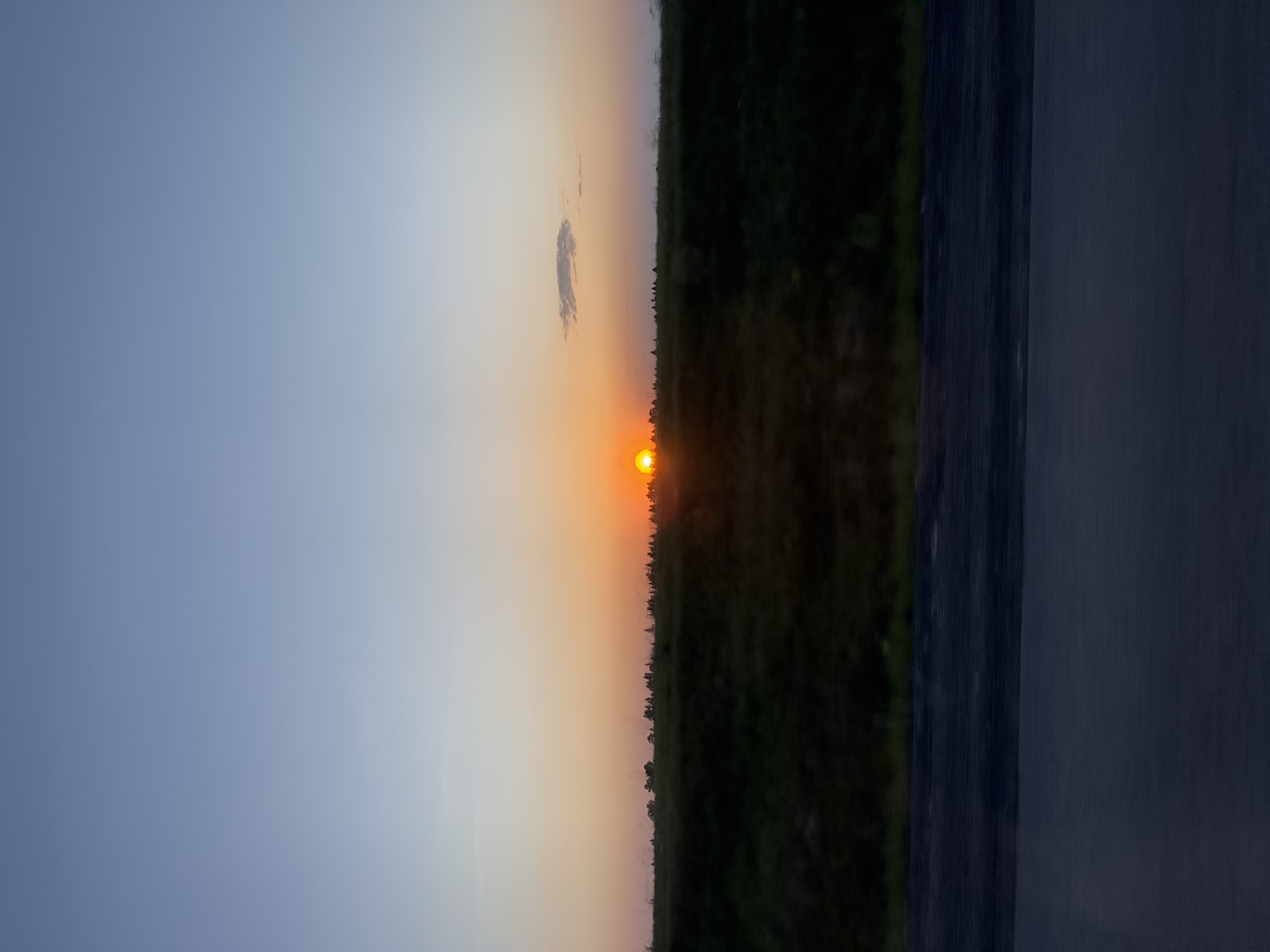 The image shows a sunset at Kamennoye, Russia. The sun is setting behind a treeline, casting an orange glow on the sky. To the left of the treeline, the sky is a light blue. There is a small cloud above the sun. A dark surface, possibly a road or wall, is present on the right of the image.
