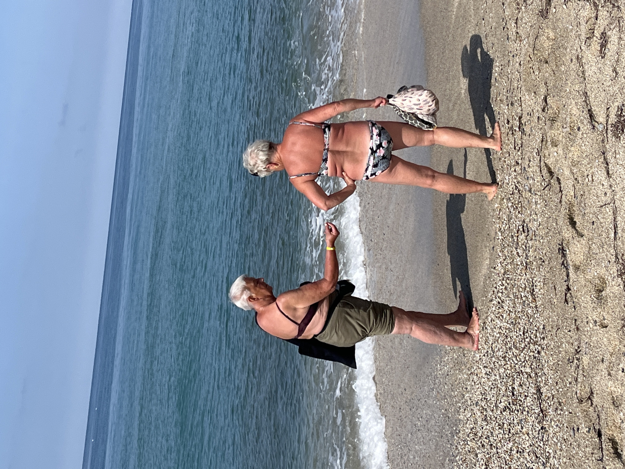 Two older women are standing on a sandy beach near the shoreline. The woman closer to the water is wearing khaki shorts and a dark-colored halter top and is facing the other woman with her arm raised. The other woman is wearing a two-piece swimsuit with a floral print and is holding a patterned tote bag. The scene is likely in Alanya, Turkey, given the beach setting and the presence of the Mediterranean Sea. The sky is slightly overcast. The beach is composed of light-colored sand mixed with small shells and some dark seaweed. The water is a muted blue color. The image appears to be captured during daylight hours.
