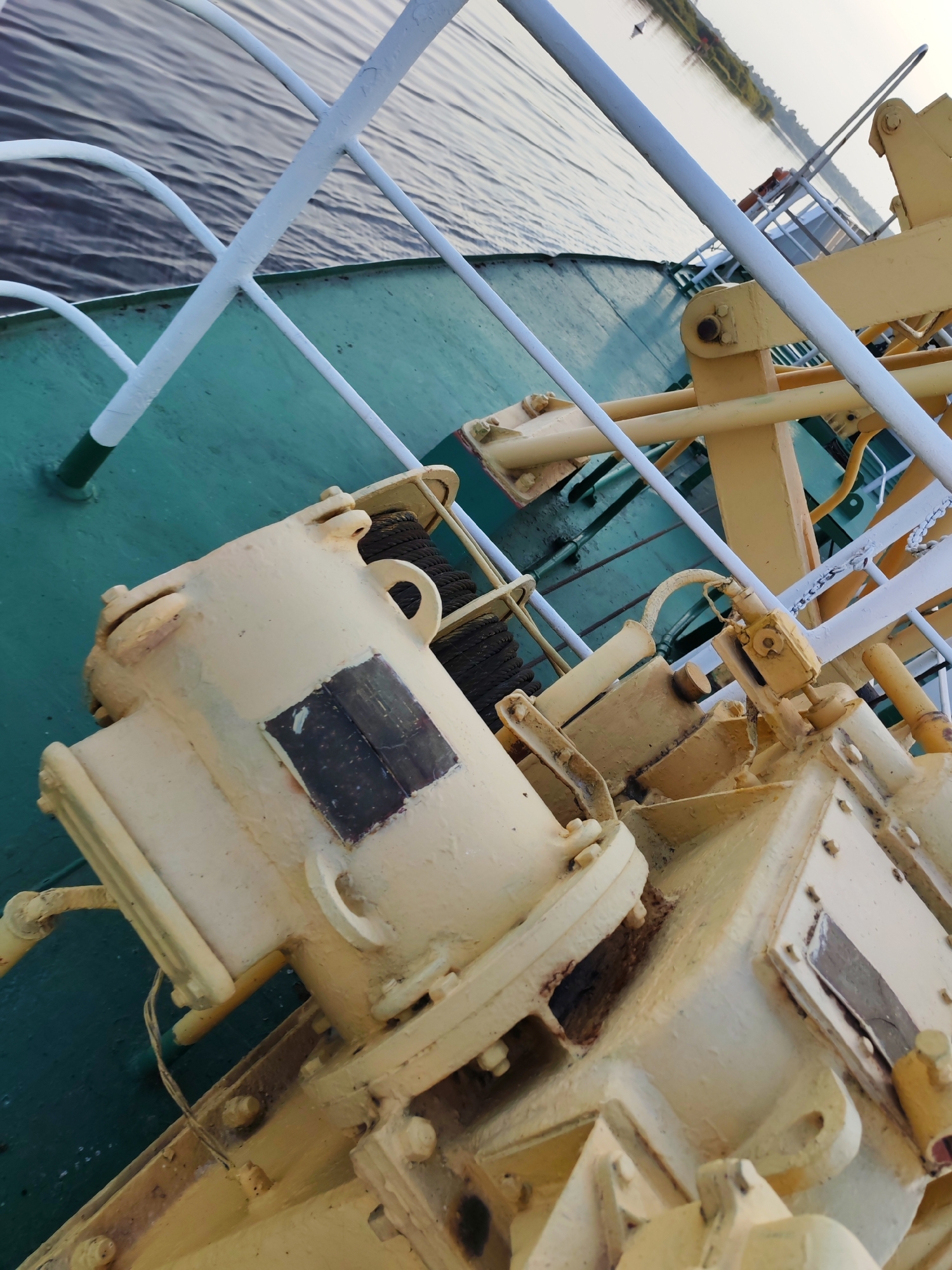The image depicts a close-up, angled view of machinery on the deck of a vessel, likely a ferry or cargo ship, on a body of water. The deck itself is painted a teal green. Several pieces of heavy, yellow-painted machinery are prominent, including what appears to be a winch or anchor mechanism with thick ropes coiled on it. Metal railings, painted white, are visible along the edge of the deck, leading towards the water. The water is calm and reflects the overcast sky. In the background, a thin strip of land with vegetation and a structure are discernible along the far bank of the waterway. The scene suggests a working vessel, possibly docked or underway, in an outdoor industrial or maritime environment. The location is identified as Sheksna, Russia.