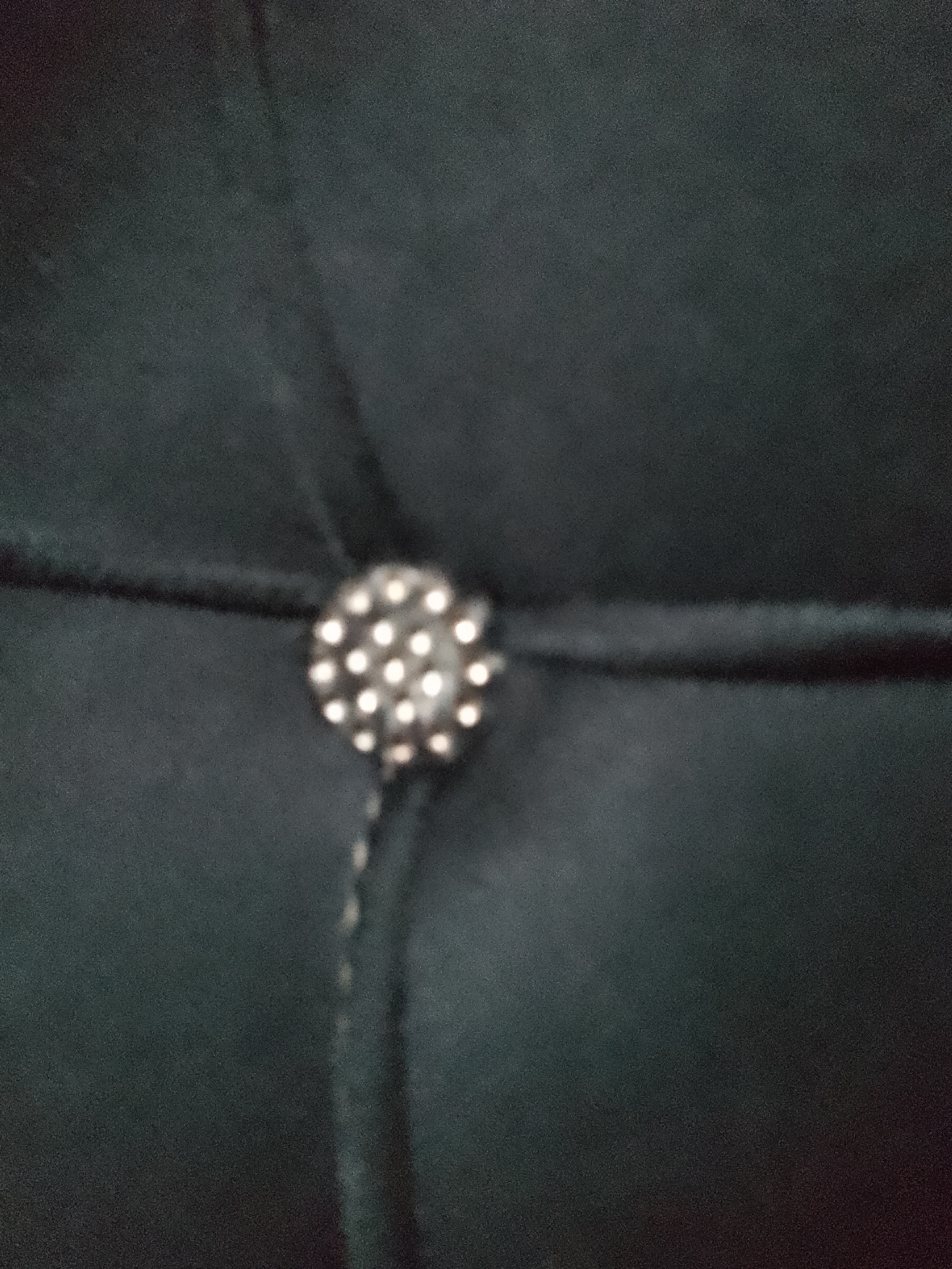 The image shows a close-up of a dark, textured surface, possibly fabric. In the center of the frame, a circular embellishment is visible. This embellishment appears to be a button or decorative stud, silver in color, with multiple small, circular indentations or studs arranged in a radial pattern. Two dark lines, likely stitching or a cord, are visible crossing horizontally and vertically through the center of the embellishment, securing it to the surface. The background is uniformly dark and shows subtle folds or creases. There is no information available to associate this image with Jauro Yinu, Nigeria.