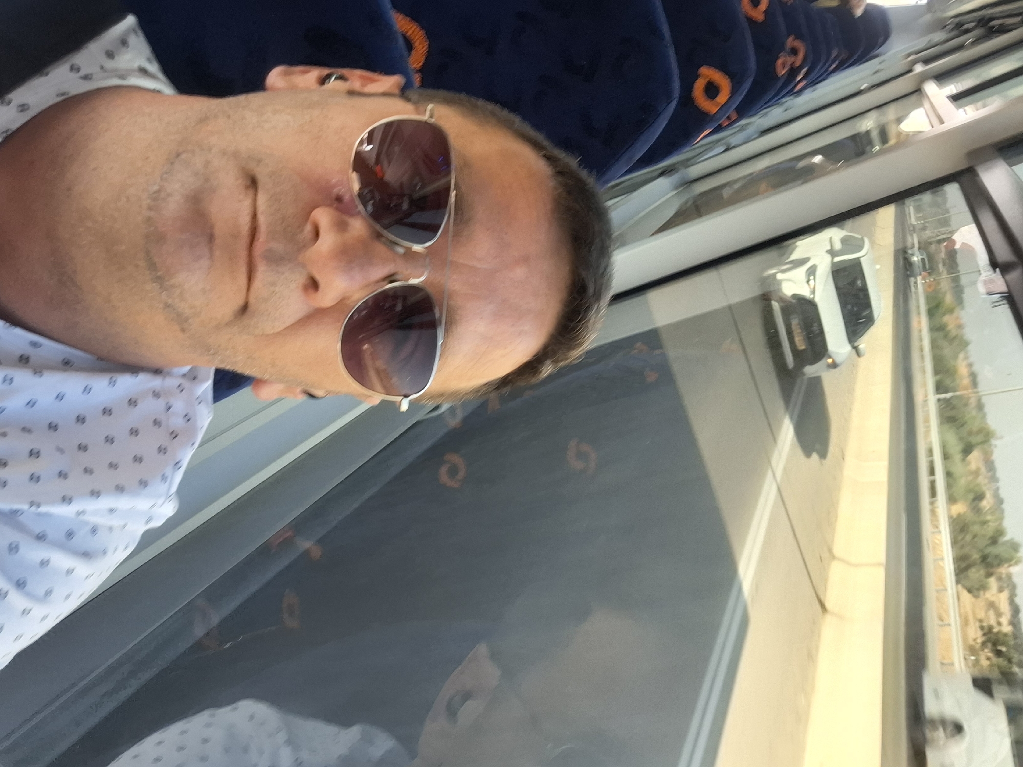 A selfie taken from inside a bus shows a middle-aged man wearing sunglasses and a white shirt with a black polka dot pattern. He is seated and has an earbud in his left ear. Through the bus window, a white car is visible on a highway, and beyond that, a landscape with sparse vegetation and a distant structure. The bus seats, upholstered in dark blue fabric with an orange symbol, are visible in the foreground and to the left of the man. The scene appears to be within the Bnei Shimon Regional Council in Israel, based on the highway and landscape.
