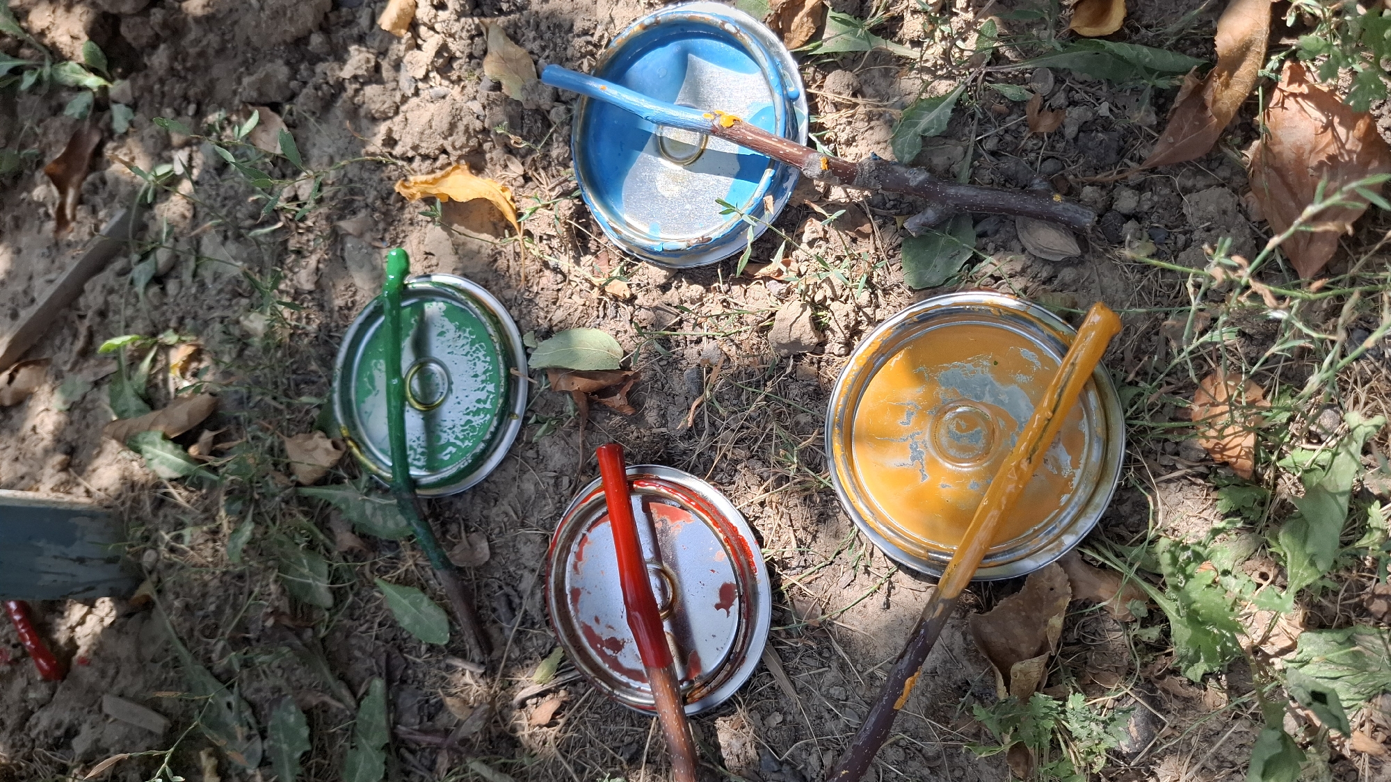 Four open tin lids, each containing a different colored paint, are laid out on the ground in a dirt and grass area. A green-painted stick lies on top of the green paint-filled lid. A red-painted stick rests on the red paint-filled lid. A blue-painted stick is on the blue paint-filled lid. An orange-painted stick rests on the orange paint-filled lid. The area appears to be outdoors.