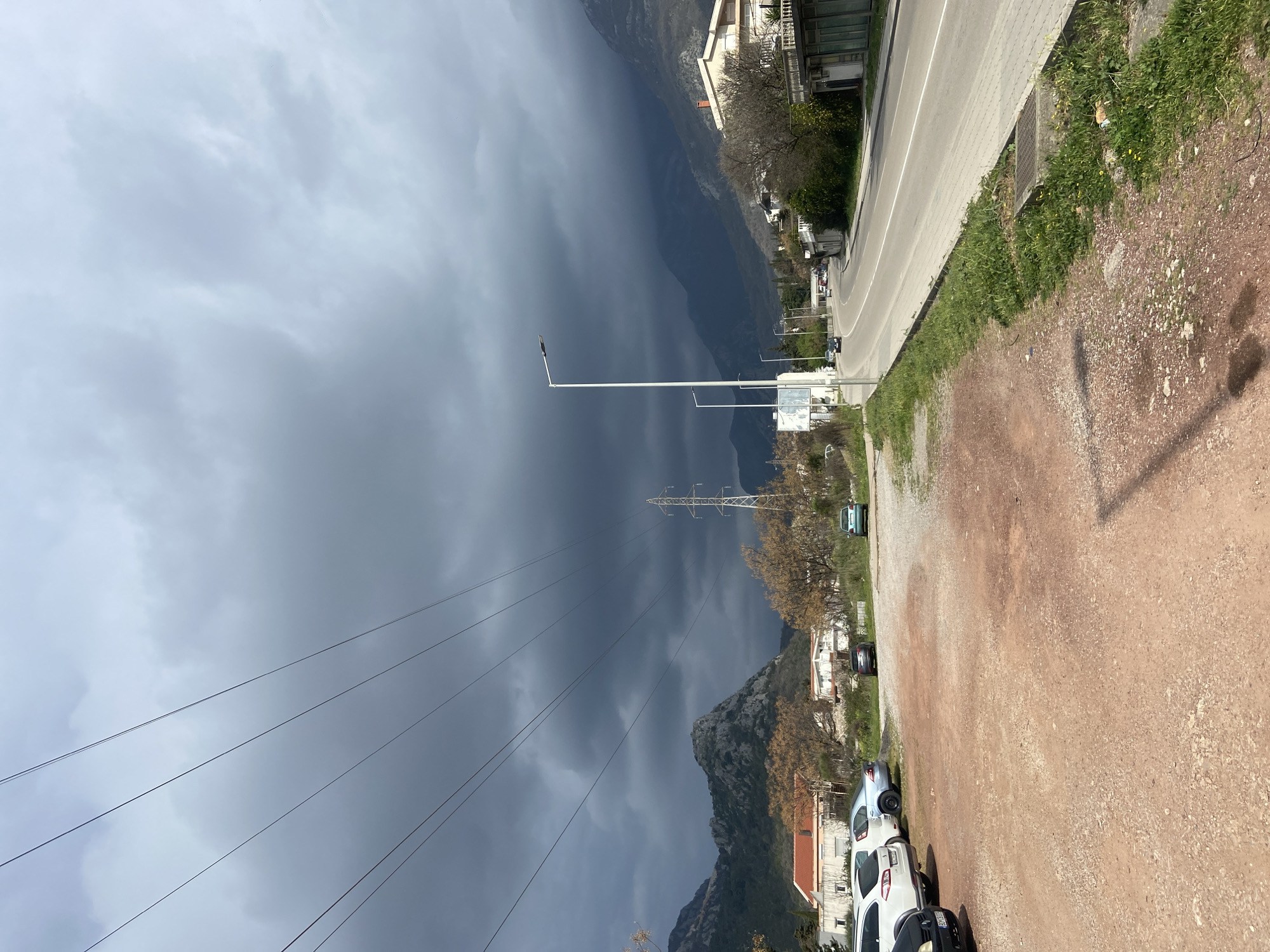 The image depicts a roadside scene in Sutomore, Montenegro, under a heavily overcast and dark sky. The foreground on the left consists of an unpaved, reddish-brown dirt and gravel parking area where five vehicles are parked: a dark front portion of a car, a white station wagon, a silver sedan, a dark gray sedan, and a light blue/green compact car.
Adjacent to the parking area on the right is a narrow strip of green grass and weeds, containing some small yellow flowers, followed by a light gray paved sidewalk. To the right of the sidewalk is a two-lane asphalt road with a solid white line separating traffic directions and a broken white line on the shoulder. Two modern white streetlights with dark fixtures are positioned along the right side of the road. Two dark vehicles are visible driving away in the distance on the road.
In the midground, beyond the parking area, are several residential or commercial buildings with light-colored facades and terracotta roofs. Deciduous trees with sparse, bare branches are scattered throughout the scene. A prominent metal electricity pylon stands in the center-left midground. Multiple overhead power lines extend horizontally across the upper portion of the frame, originating from or passing near the pylon.
The background features dark, rugged mountains, particularly on the left and right, obscured by the low-hanging, dark gray storm clouds that dominate the upper sky. The overall light condition is dim due to the extensive cloud cover. No individuals are visibly present or engaged in specific actions.