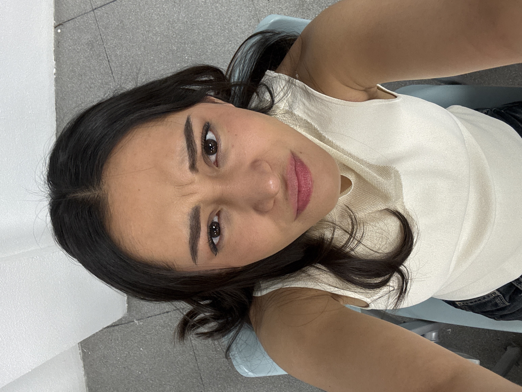 An overhead-angle image depicts a single adult female subject seated indoors. The woman, with dark brown hair and medium complexion, looks directly into the camera. Her facial expression is characterized by furrowed eyebrows and downturned lips, forming a grimace or pout. She is attired in a cream-colored, ribbed sleeveless top and dark-colored trousers. A light blue chair is partially visible beneath her, serving as her seat. The immediate environment consists of a light grey tiled floor and a plain white wall or ceiling in the background, suggesting an unadorned interior space. The specific location within Puebla, Mexico, is not discernible from the visual information presented.