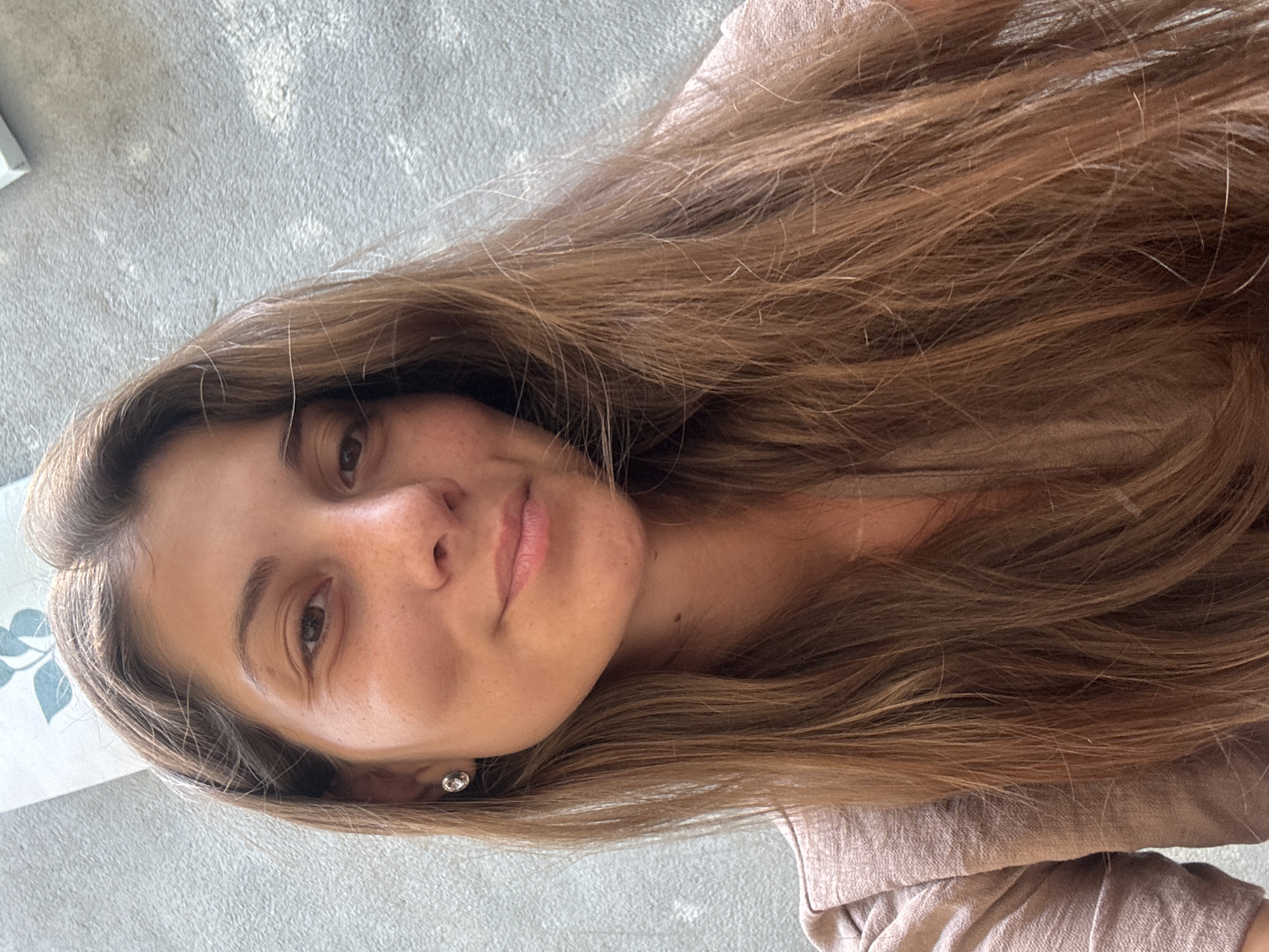 A woman with long, light brown hair, fair skin, and freckles is facing the camera. She is wearing a light brown shirt. The background is a light gray surface. The location is impossible to determine from the image.