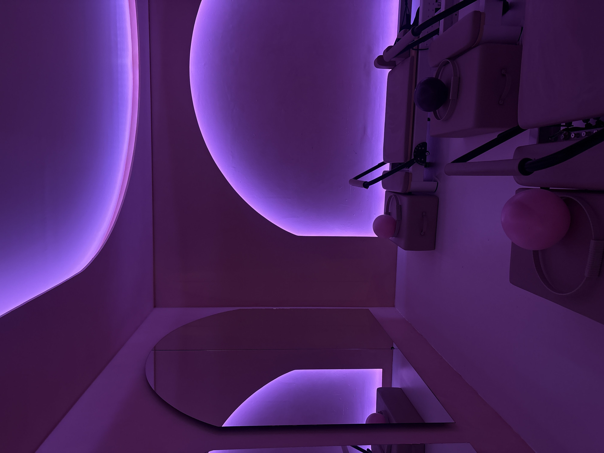 A rotated interior view of a pilates studio in Mexico City, Mexico, lit with purple lights. The walls are white and decorated with mirrors. Pilates equipment is mounted on the walls.