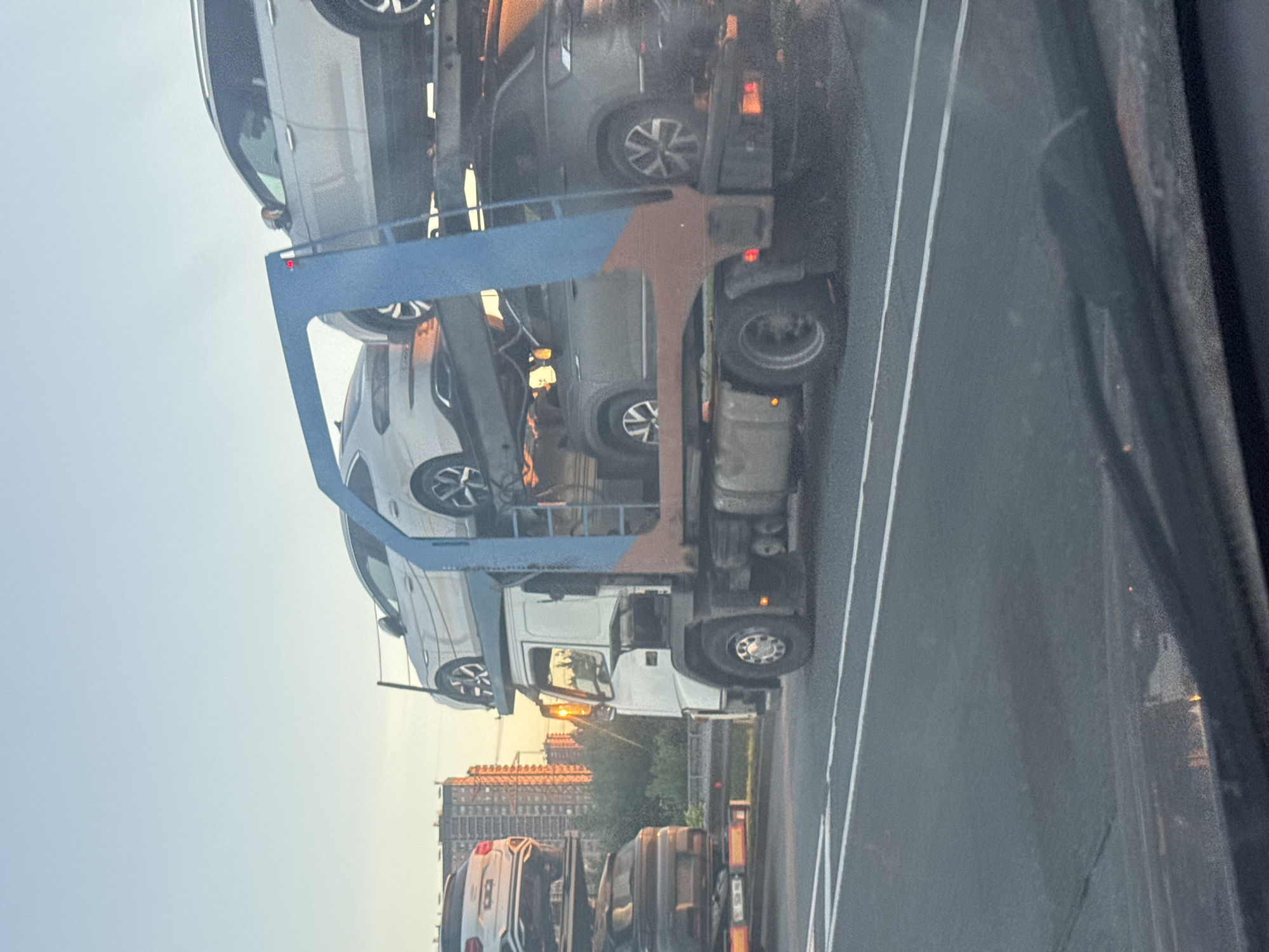 An auto transport truck is shown on a highway with a golden sky suggesting either sunrise or sunset. The truck is carrying multiple cars on its upper decks. One car, a white sedan, is clearly visible on the top deck. Below it, a dark colored SUV is visible on a lower deck. In the background, a cluster of tall buildings, likely apartment blocks, are illuminated by the setting or rising sun. The truck appears to be in transit, likely on a road in or near Poroshkino, Russia, given the architectural style of the buildings. The perspective of the image is from inside another vehicle, looking out at the truck.