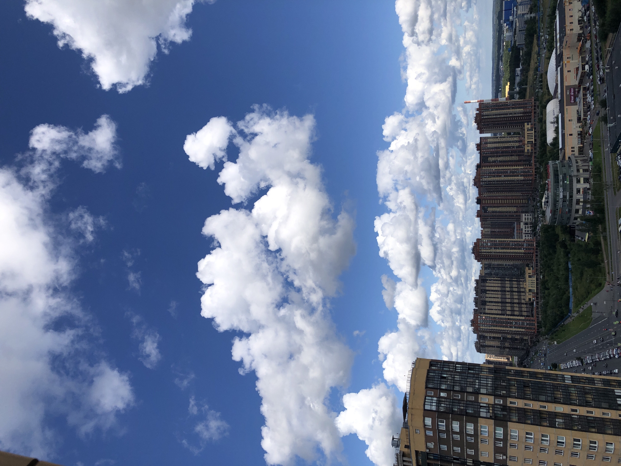 This is a high-angle, vertical shot of a cityscape with a bright blue sky filled with fluffy white clouds. In the foreground on the left is a tall, modern, multi-story building with a tan and brown facade, featuring many windows. To the right of this building is a wide, straight road lined with parked cars. Beyond the road, a row of similarly tall, modern apartment buildings stretches across the middle ground, displaying a variety of architectural details and colors, primarily brown and beige. In front of these buildings, there is a patch of green trees, and further in the distance, more buildings and a large red and white striped smokestack are visible. The overall environment suggests an urban residential area on a clear, cloudy day.