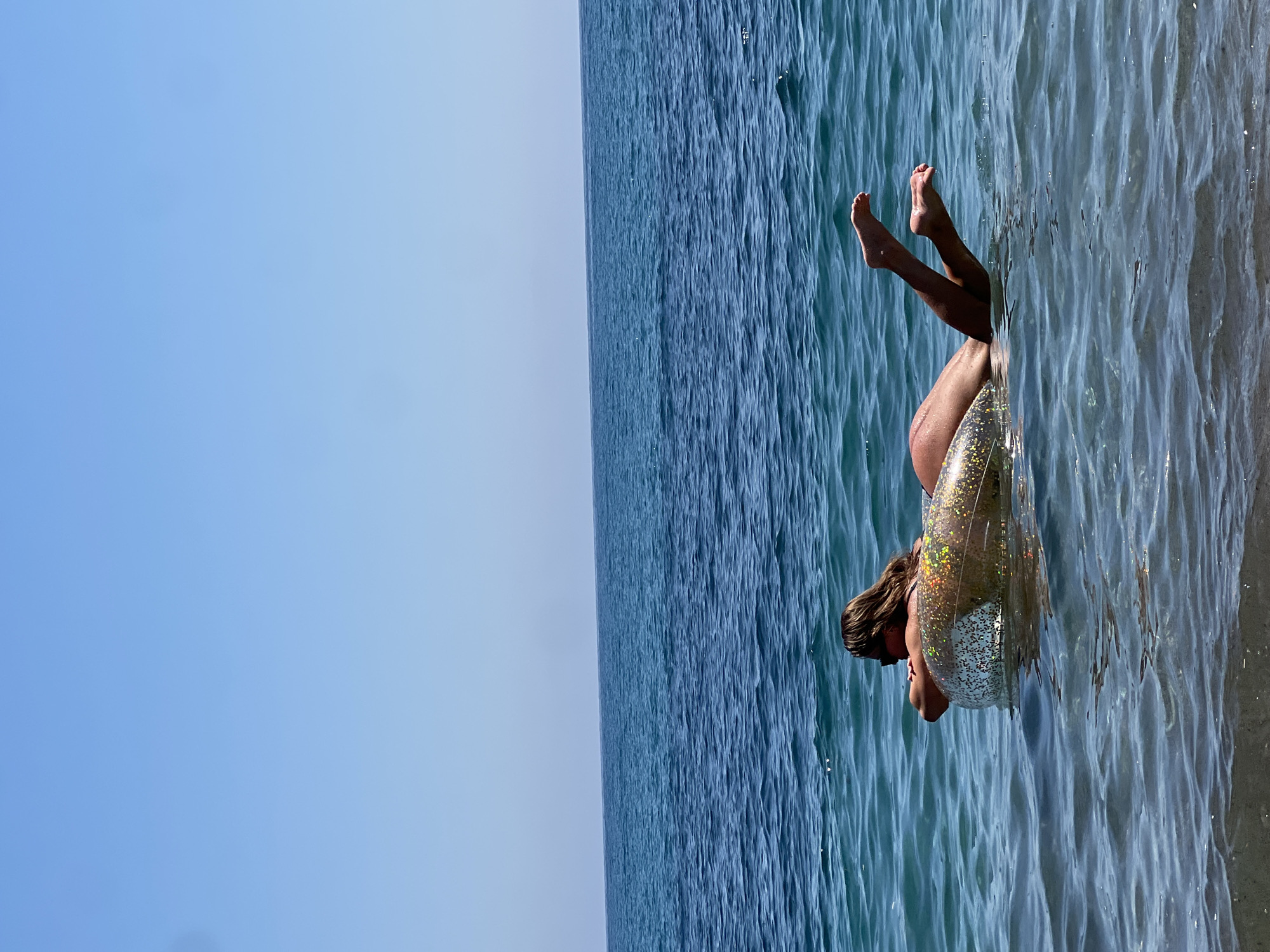 The image, oriented horizontally, depicts a person relaxing in the sea under a clear blue sky. The scene is set in Prijevor, Montenegro. The individual, likely female, is partially submerged in calm, rippling blue-green water, supported by a transparent inflatable ring filled with glittering specks. Her lower body, including legs and feet, is raised above the water, while her head and shoulders are visible on the other side of the ring. Her hair is dark brown, and she appears to be wearing a swimsuit. The water shows gentle waves and reflections of sunlight, indicating a bright, sunny day. The horizon line is visible, separating the expanse of the tranquil sea from the uniformly clear, light blue sky above. The overall impression is one of leisure and enjoyment in a serene, natural environment. No text is visible in the image.