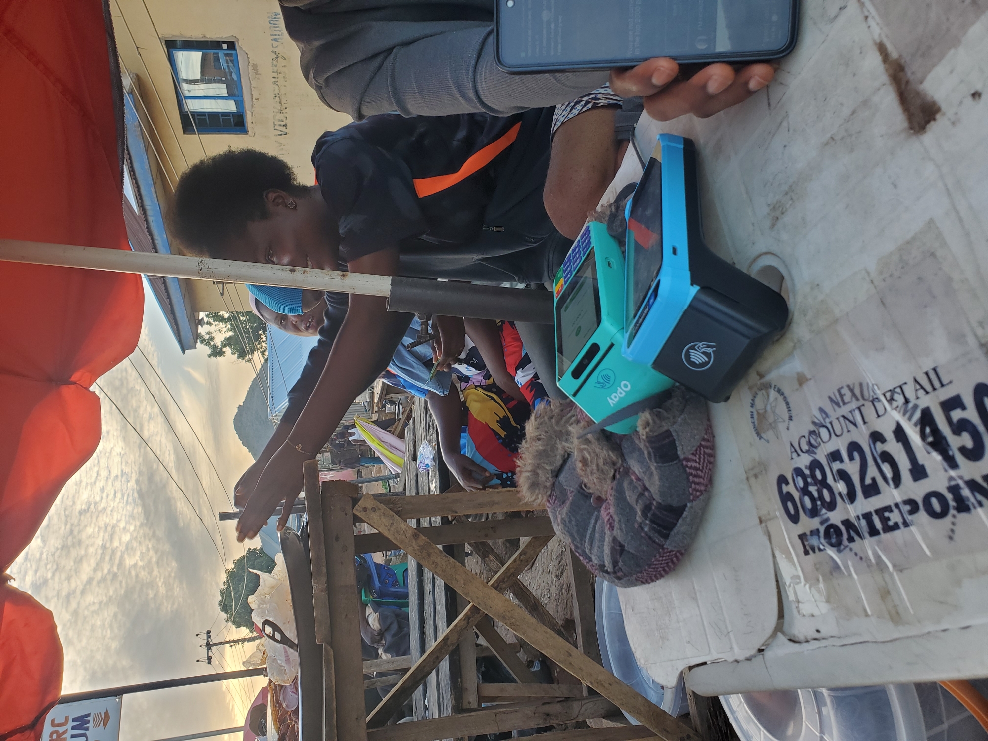 In Gwong, Nigeria, at an outdoor market stall, a young woman with dark, curly hair and wearing a black jacket with an orange stripe on the sleeve is captured in profile. She is looking to her right, her hands raised and slightly blurred, suggesting movement or an interaction. Two blue and green point-of-sale devices are visible on a table in front of her, one with an OPay logo. A patterned cloth is also on the table, to the left of the devices. In the background, a building with blue-trimmed windows and text that reads "VICKEY REALM SALON" is visible, along with electrical poles and a cloudy sky. The scene appears to be set during the late afternoon or early morning, given the lighting conditions.