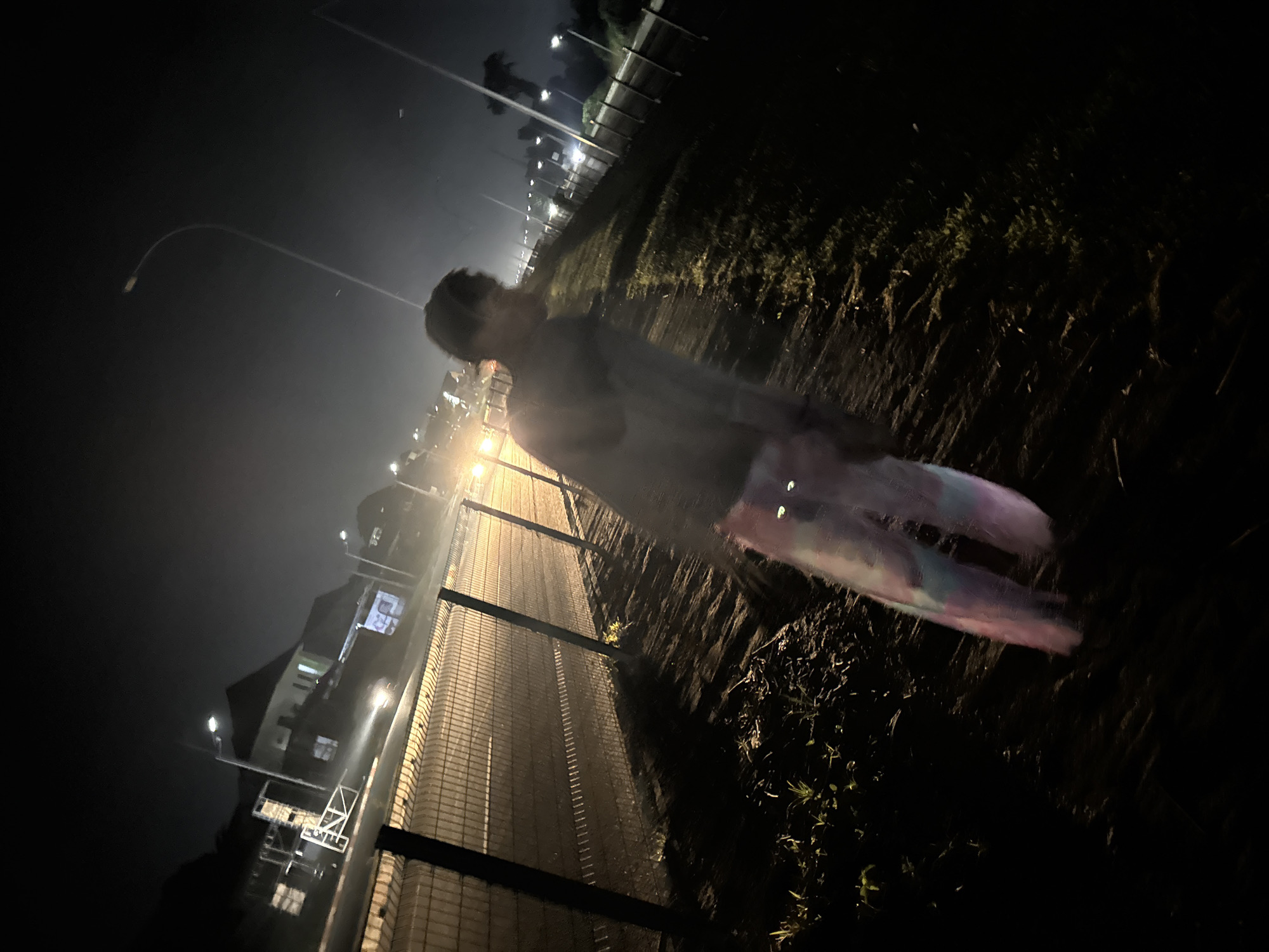 A person walks along a dirt path next to a chain-link fence at night in Uyo, Nigeria. The person is blurry due to motion, and their pants have a tie-dye pattern of pink, blue, and white. Streetlights illuminate the scene, casting shadows and creating a hazy glow. In the background, there are buildings and more streetlights along a road.